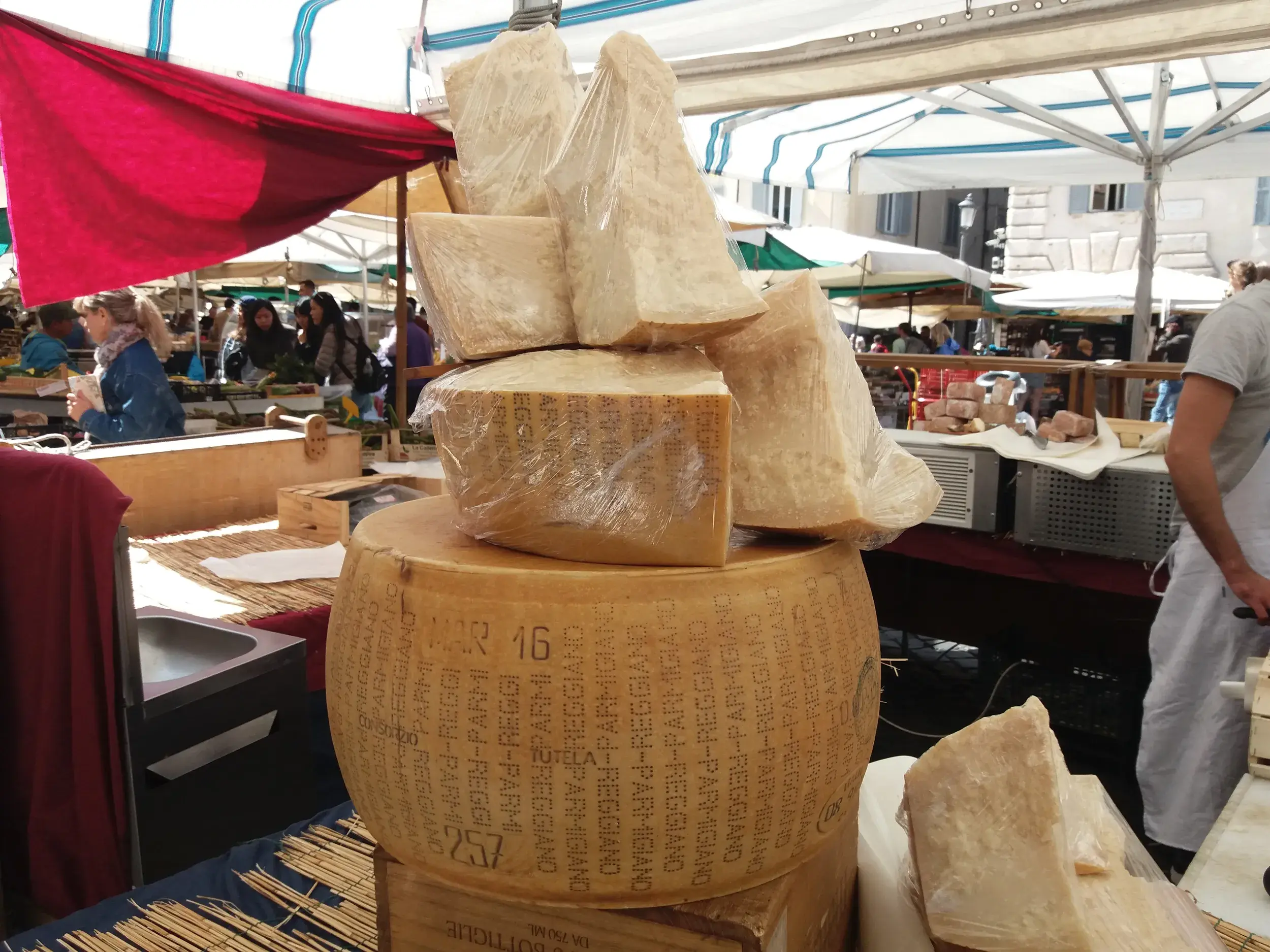 Ein großer Laib Parmesan-Käse mit aufgetürmten Stücken auf dem Stand eines belebten Wochenmarktes in Italien.