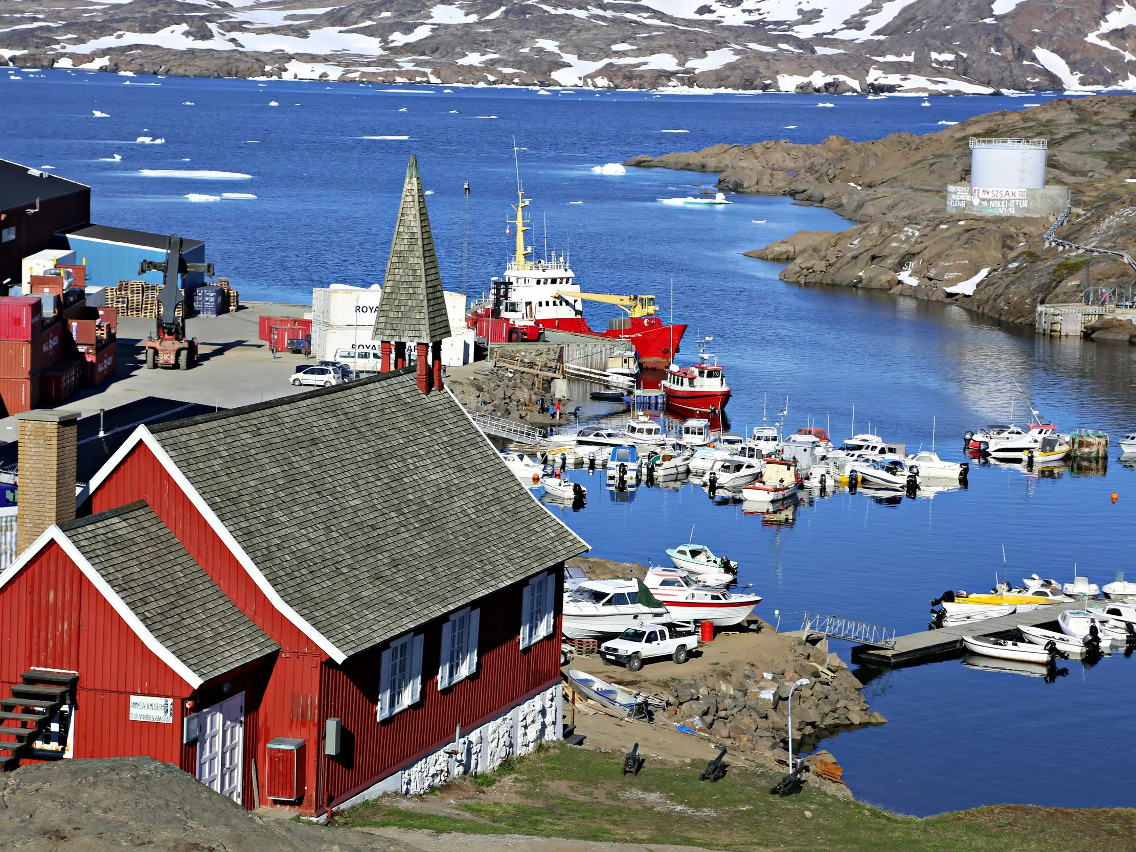 Rotes Holzhaus am Hafen von Tasiilaq in Ostgrönland. Boote und Schiffe liegen im Fjord, im Hintergrund treiben Eisberge.
