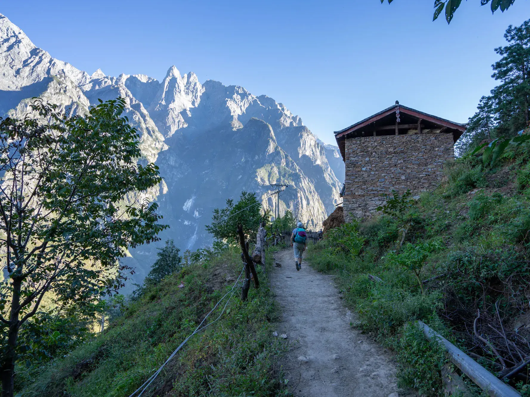 Ein Wanderer auf einem schmalen Pfad entlang eines Steinhauses bei einer Trekking-Tour in der Tigersprungschlucht in Ost-Tibet.