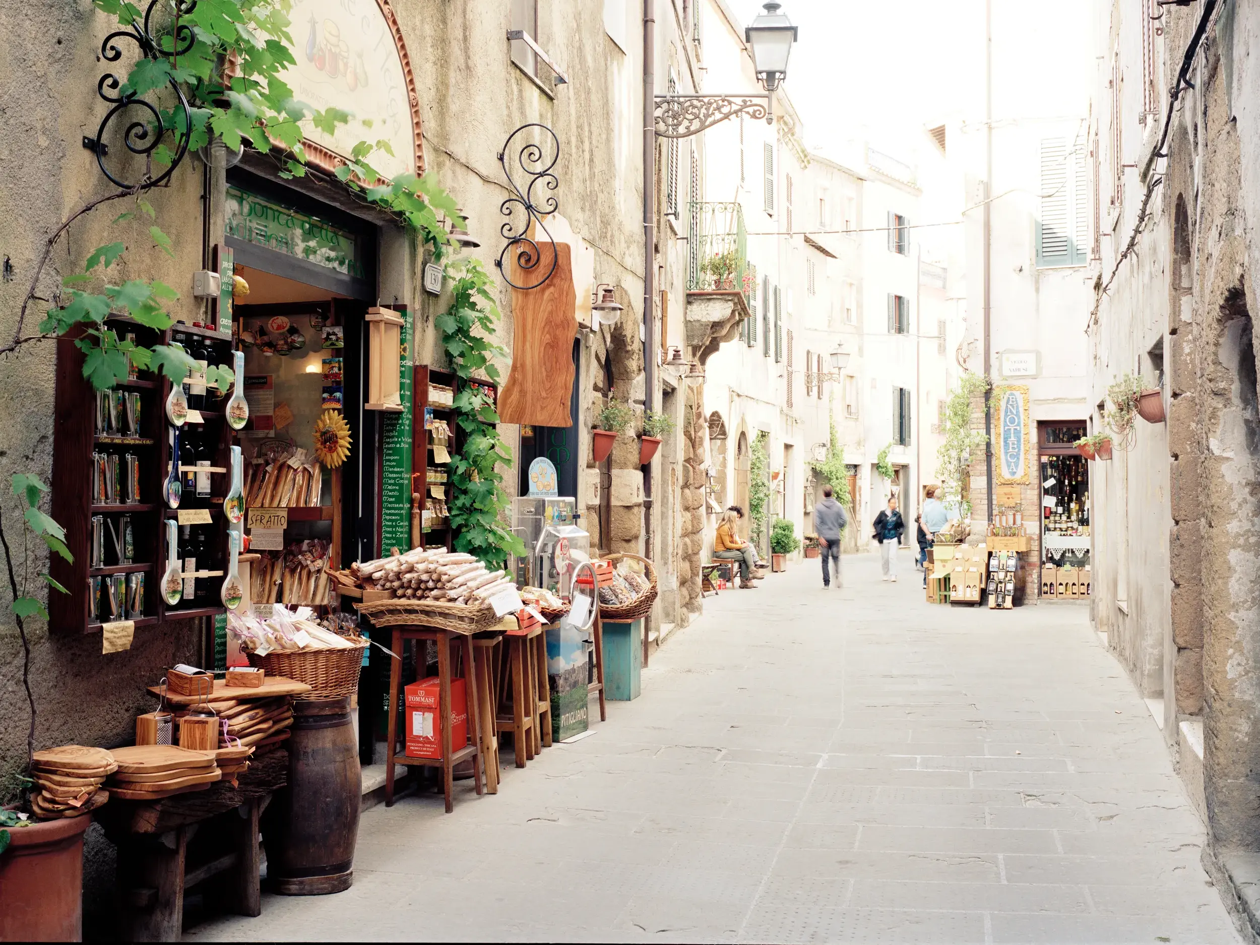Ein Geschäft für Delikatessen säumt eine enge Gasse in Massa Marittima, im Hintergrund spazieren Besucher durch die Altstadt.