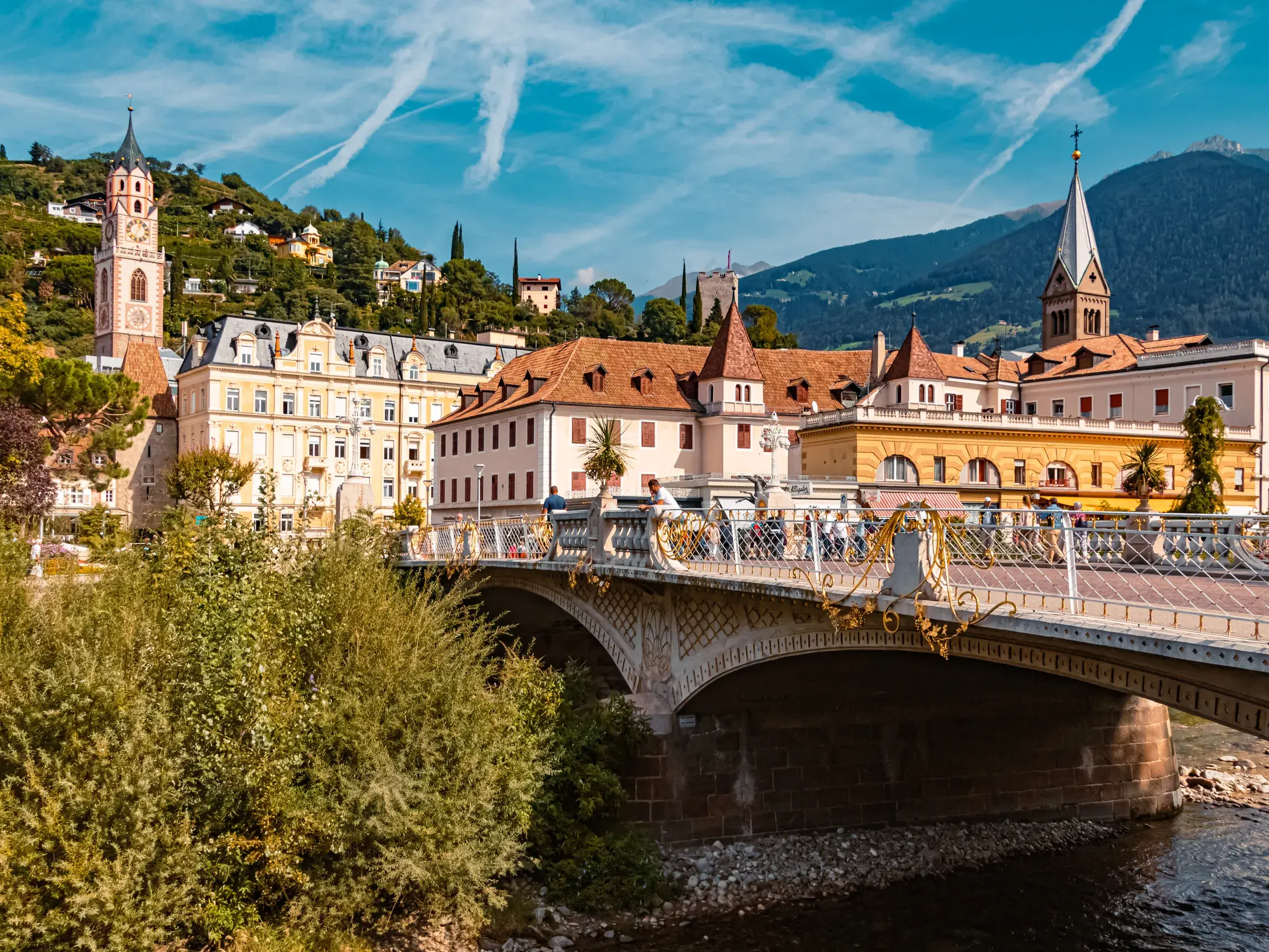 Blick über die historische Postbrücke auf die Altstadt von Meran in Südtirol bei sonnigem Wetter mit Bergen im Hintergrund.