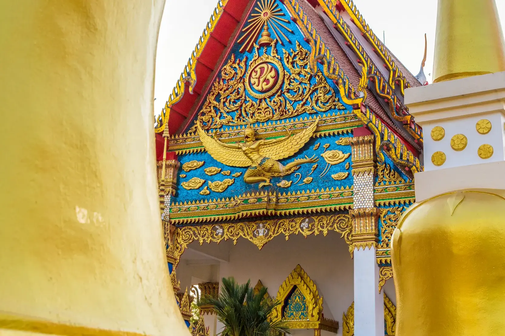 Nahaufnahme des prächtig verzierten Giebels eines Tempels in Thailand mit einer goldenen Garuda-Figur vor blauem Mosaik.