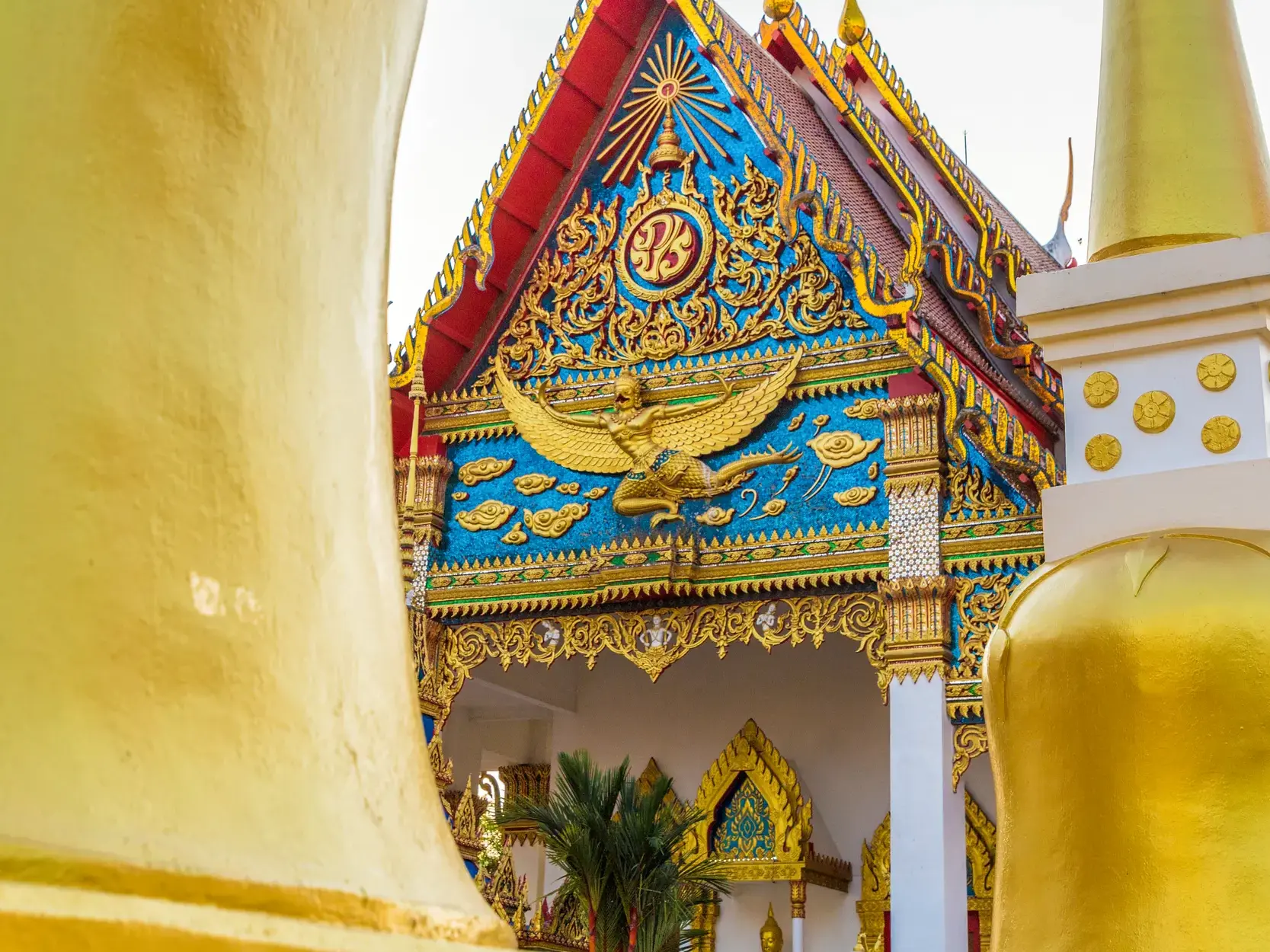 Nahaufnahme des prächtig verzierten Giebels eines Tempels in Thailand mit einer goldenen Garuda-Figur vor blauem Mosaik.