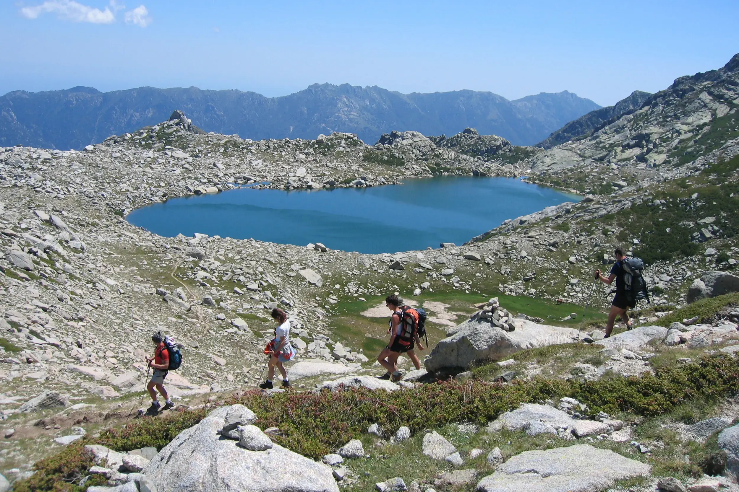 Vier Personen beim Trekking auf dem GR20 in Korsika wandern auf einem steinigen Pfad oberhalb eines blauen Gletschersees.