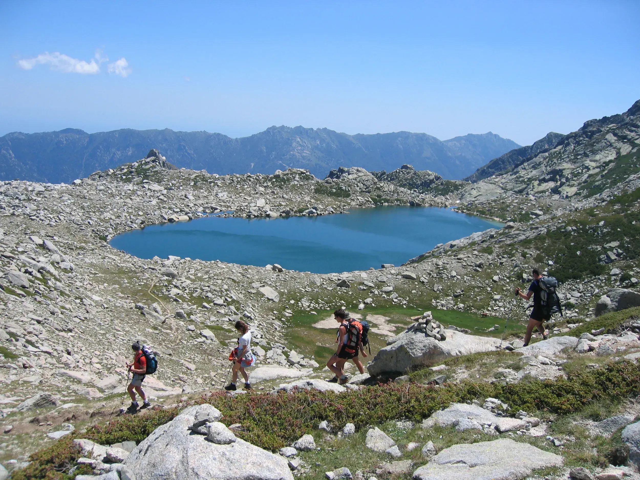 Eine Wandergruppe steigt auf dem GR20 auf Korsika ab, mit Blick auf einen blauen Gletschersee in einer felsigen Hochgebirgslandschaft.