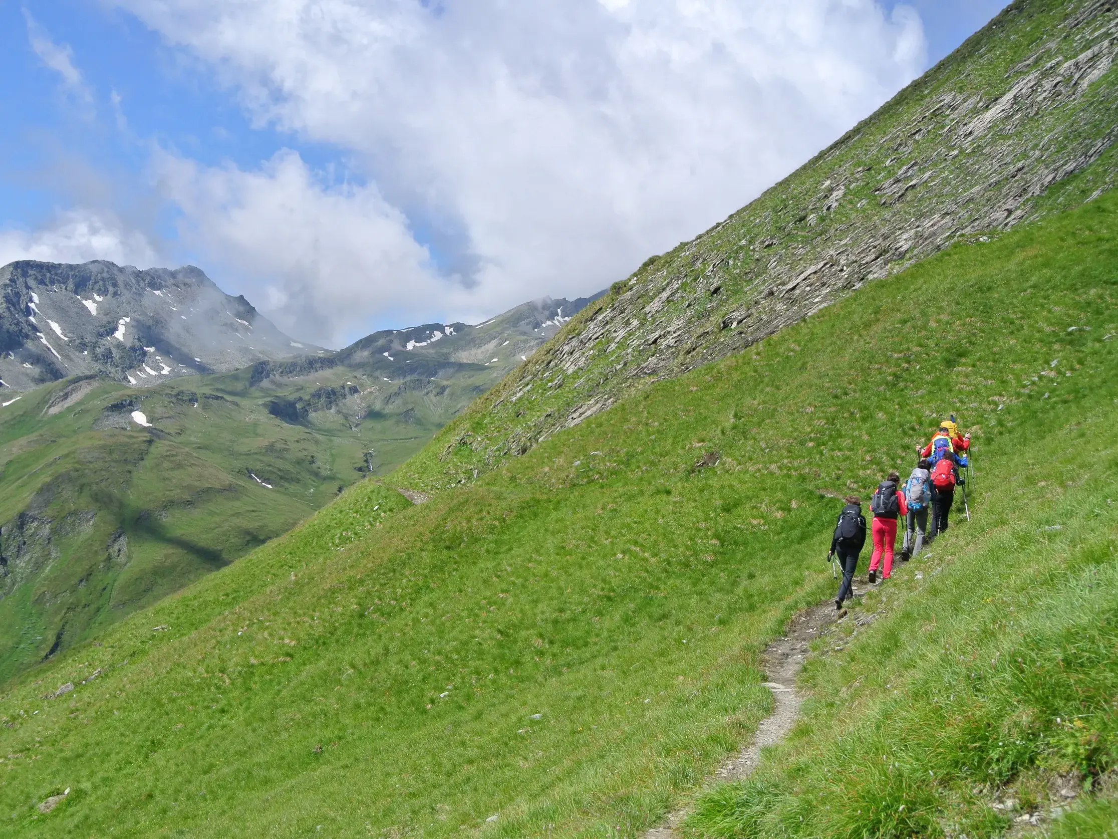 Eine Wandergruppe mit Rucksäcken auf einem schmalen Pfad an einem steilen, grünen Hang im Nationalpark Hohe Tauern.
