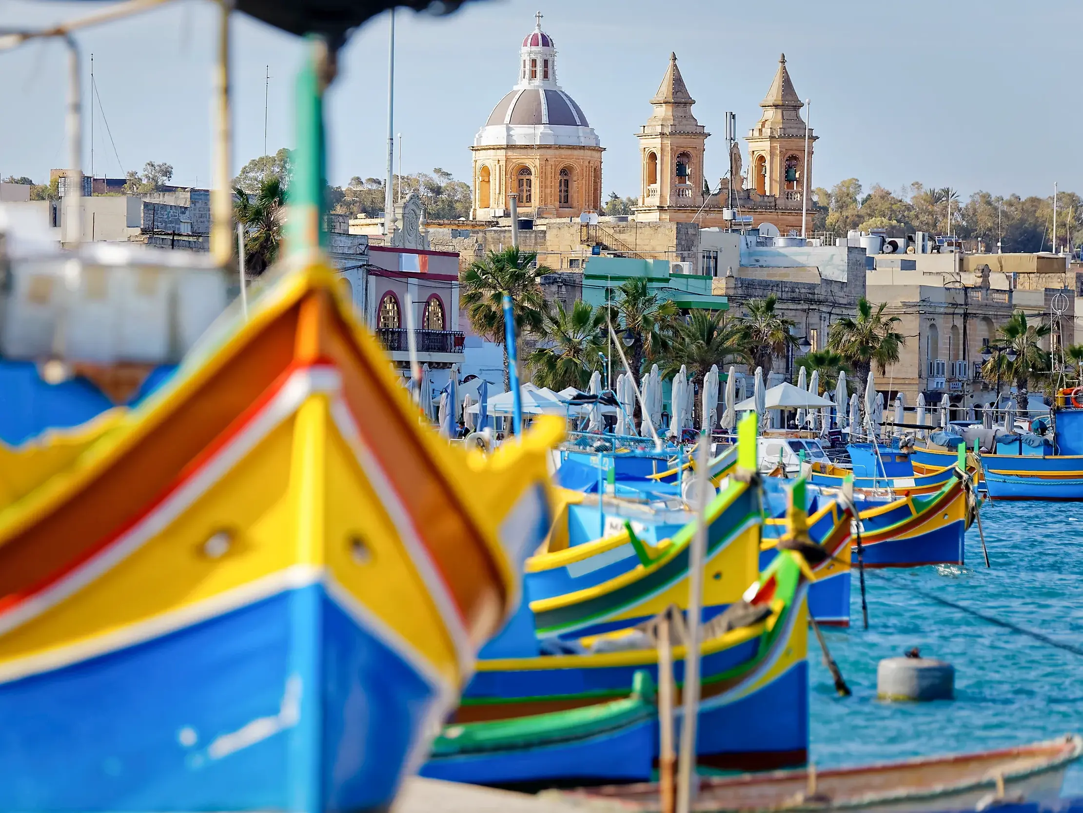 Traditionelle, bunte Fischerboote liegen im Hafen von Marsaxlokk vor der Kulisse der Stadt und einer barocken Kirche auf Malta.