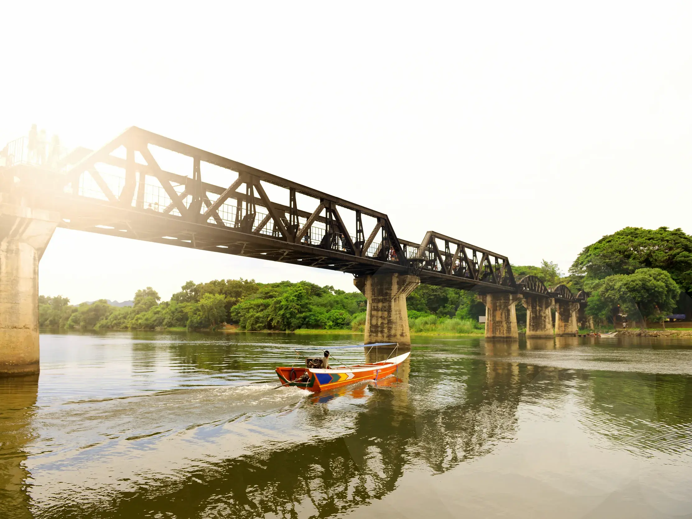 Ein buntes Langschwanzboot fährt bei Sonnenschein auf dem River Kwai unter der historischen Eisenbahnbrücke in Kanchanaburi.
