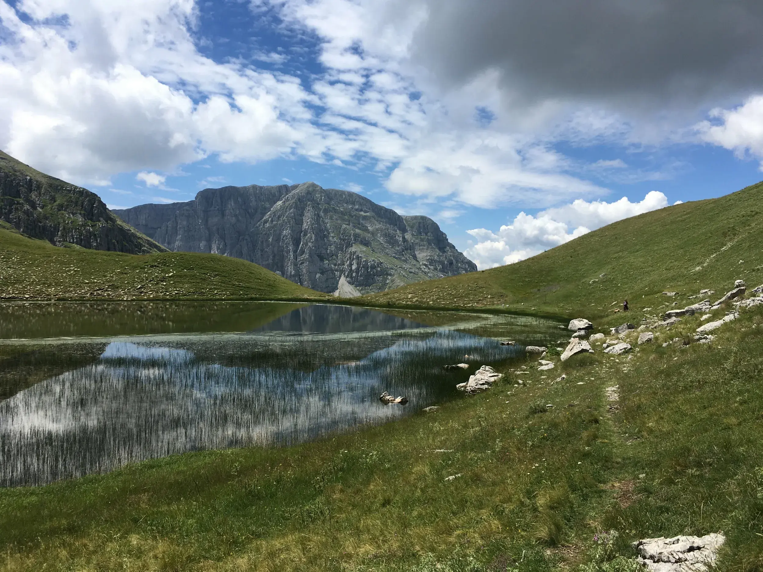 Wolken spiegeln sich im klaren Wasser des Drachensees Drakolimni, umgeben von grünen Hängen des Pindos-Gebirges in Griechenland.