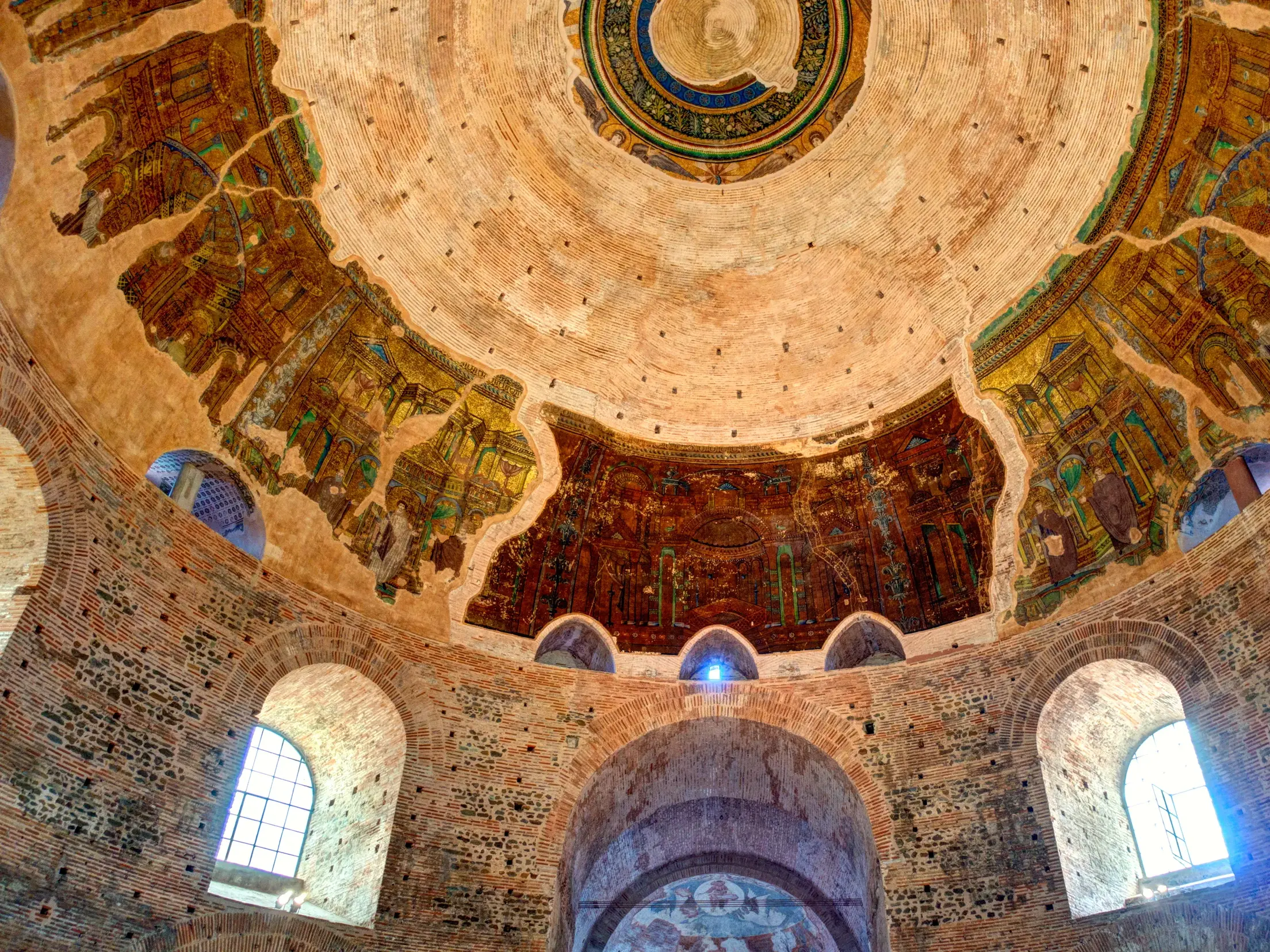 Blick nach oben in die monumentale Backsteinkuppel in Thessaloniki mit teils zerstörten, aber farbenprächtigen Mosaiken.