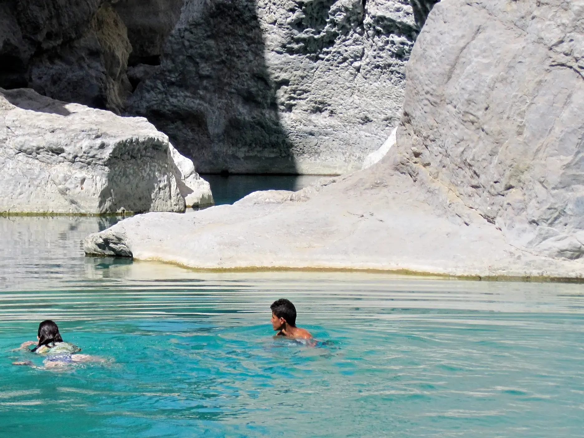 Zwei Wanderer erfrischen sich beim Schwimmen im türkisblauen Felsenpool der von hellen Felsen umgebenen Wadi Shab Schlucht.