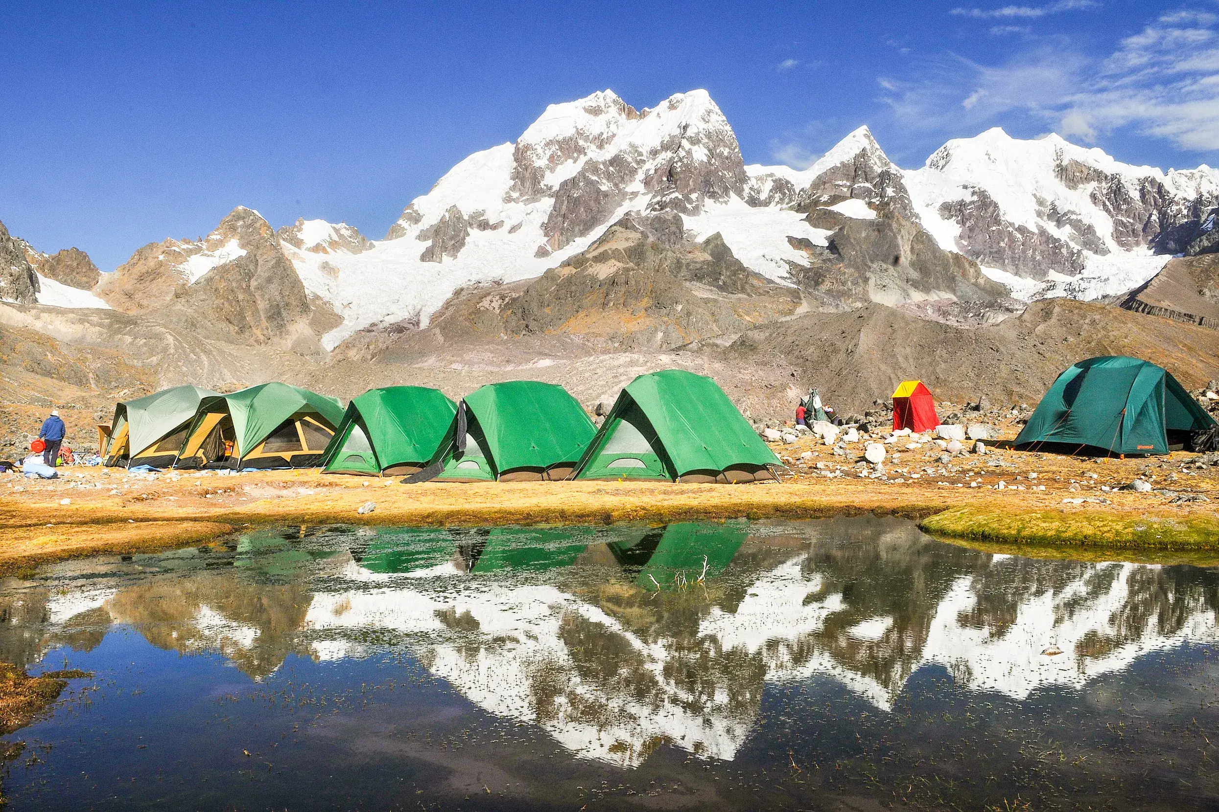 Aufgeschlagene Trekking-Zelte am Ufer eines Bergsees, in dem sich die schneebedeckten Gipfel der Cordillera Vilcanota spiegeln.