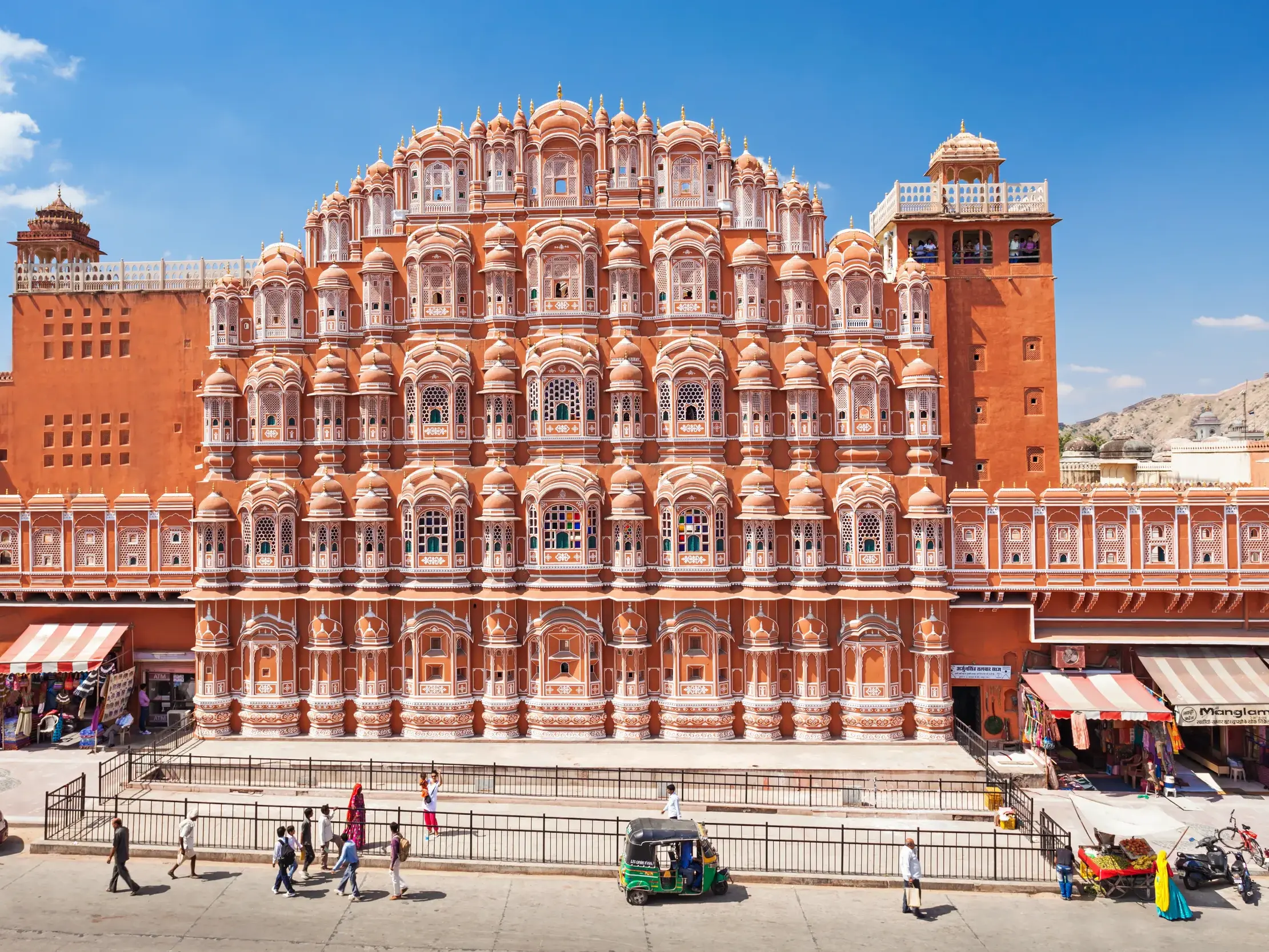Die rosa Fassade des Palastes der Winde in Jaipur mit hunderten Fenstern an einer belebten Straße mit Passanten und Rikschas.
