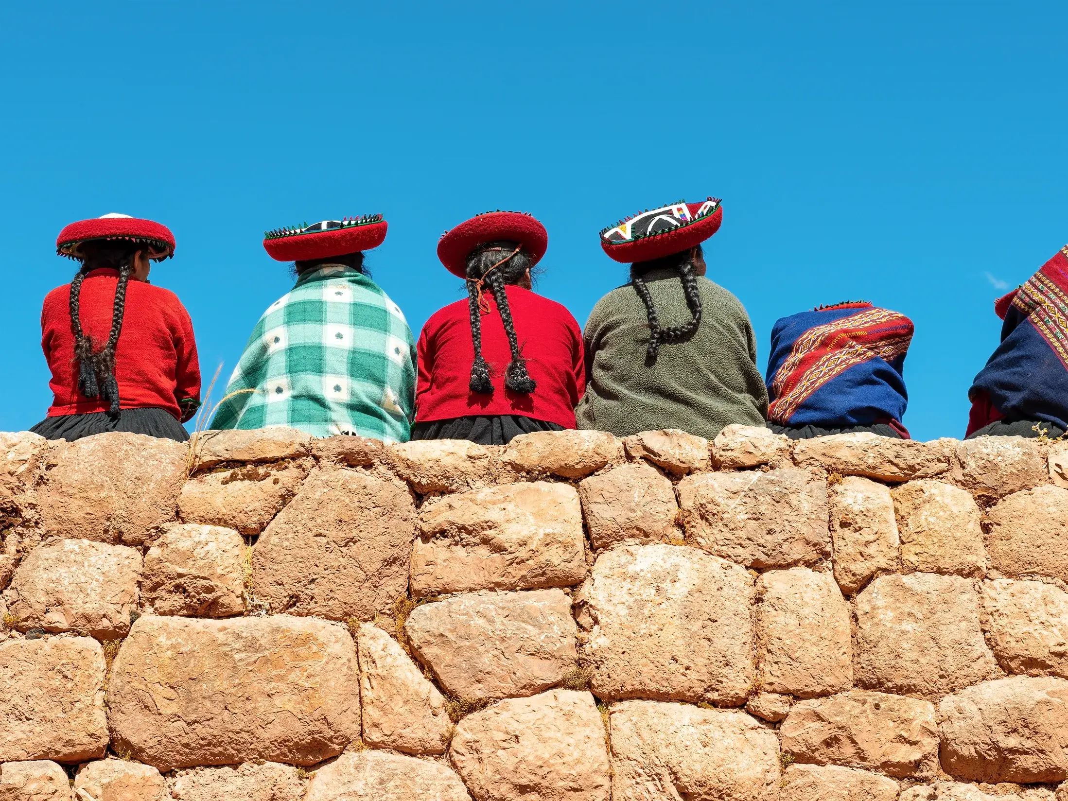 Sechs peruanische Frauen in traditioneller, farbenfroher Kleidung sitzen auf einer alten Inka-Mauer im Heiligen Tal.