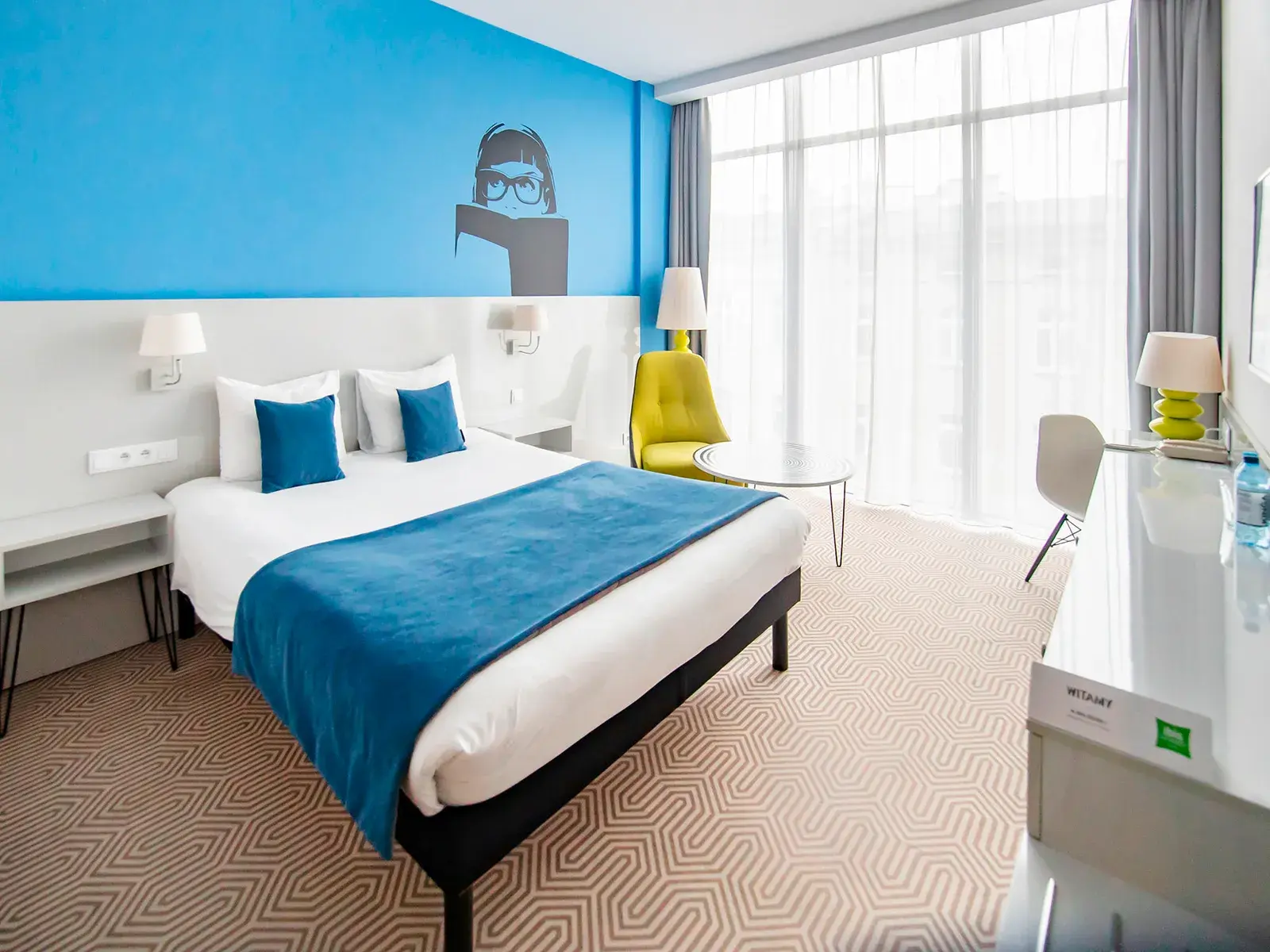 Modernes Hotelzimmer in Polen mit Doppelbett vor einer blauen Wand mit Grafik, Schreibtisch und heller Fensterfront.