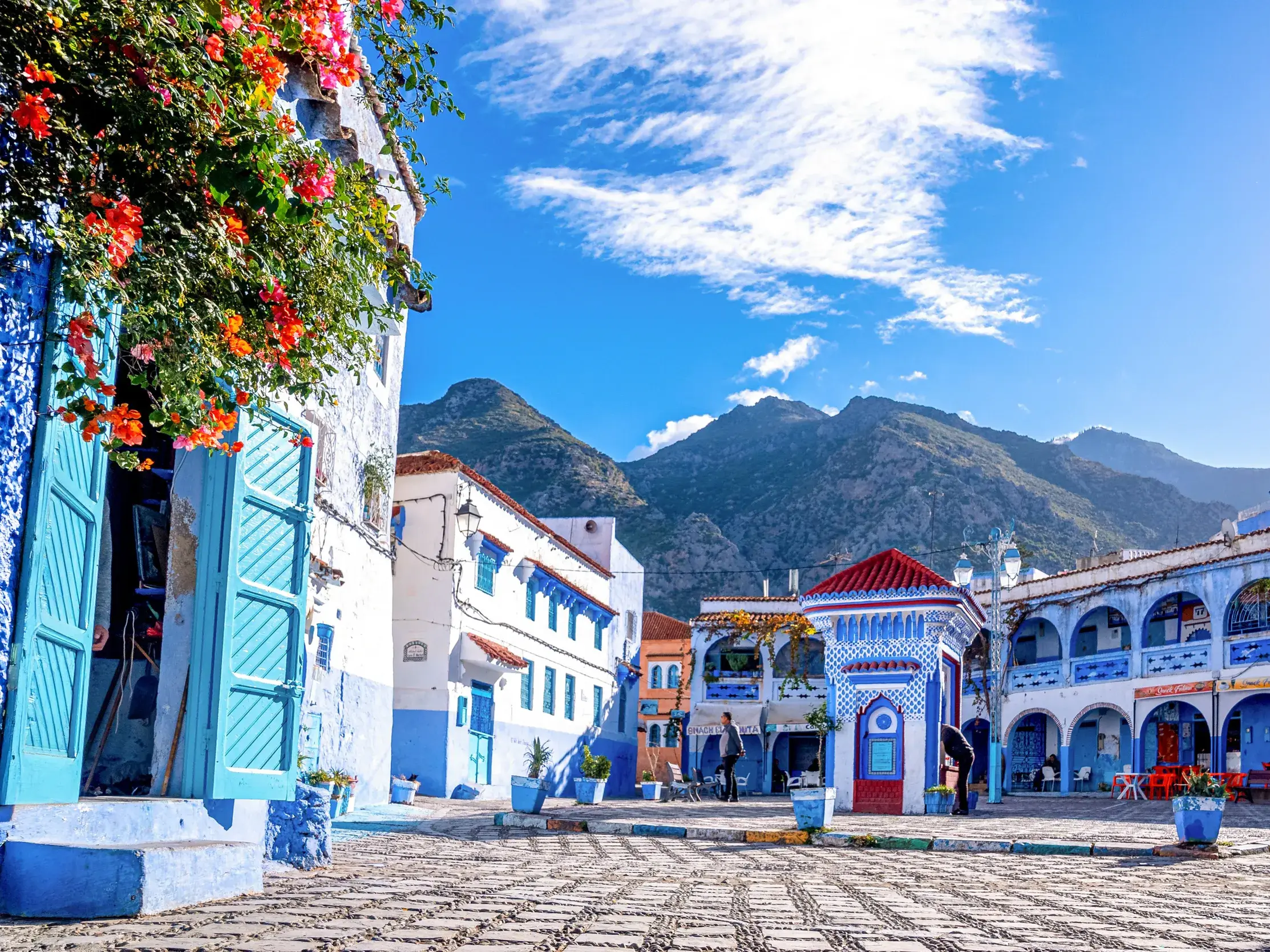 Sonniger Platz mit blauen Häusern in der Medina von Chefchaouen vor der Bergkulisse des Rif-Gebirges in Marokko.
