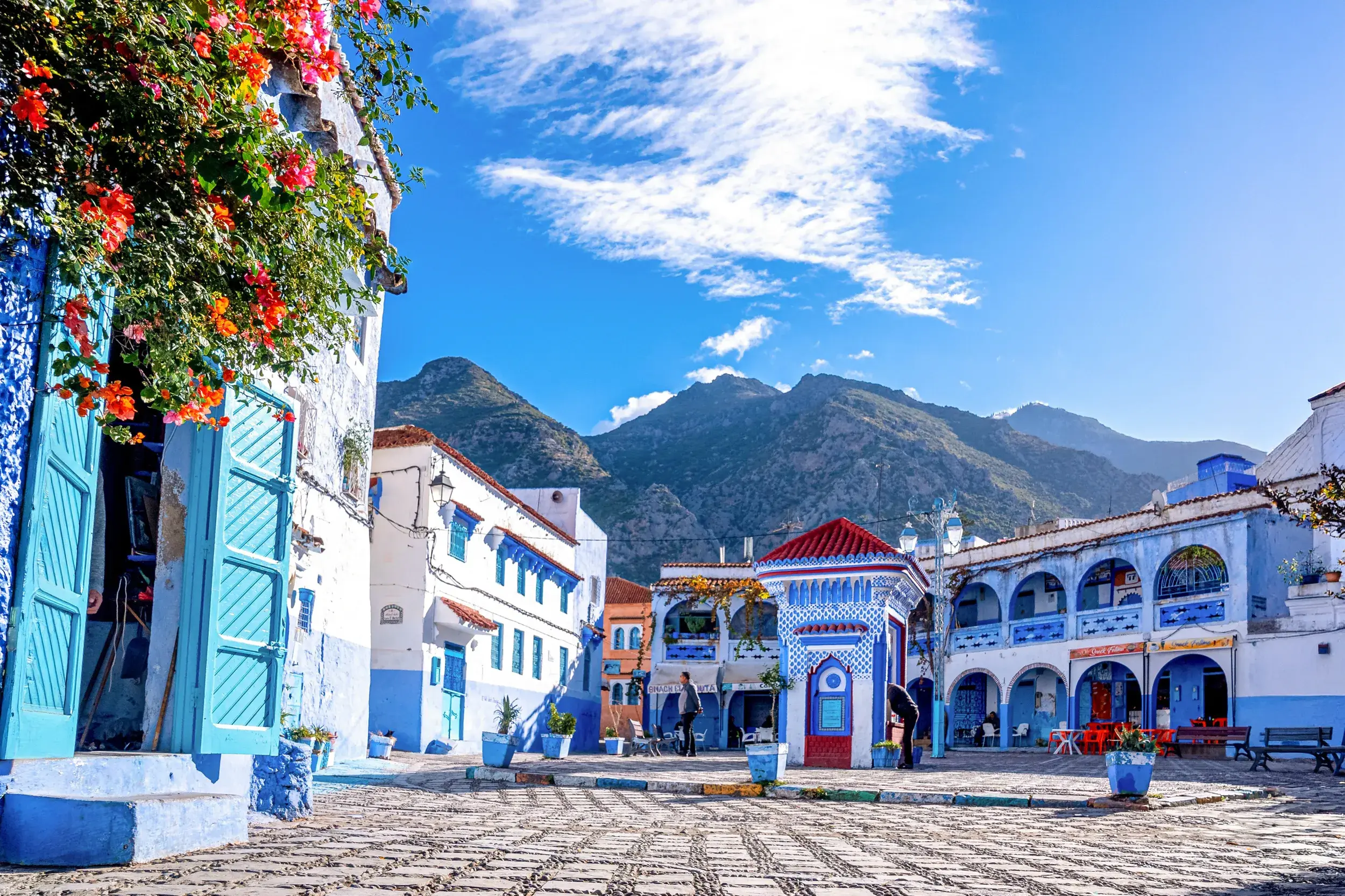 Sonniger Platz mit blauen Häusern in der Medina von Chefchaouen vor der Bergkulisse des Rif-Gebirges in Marokko.