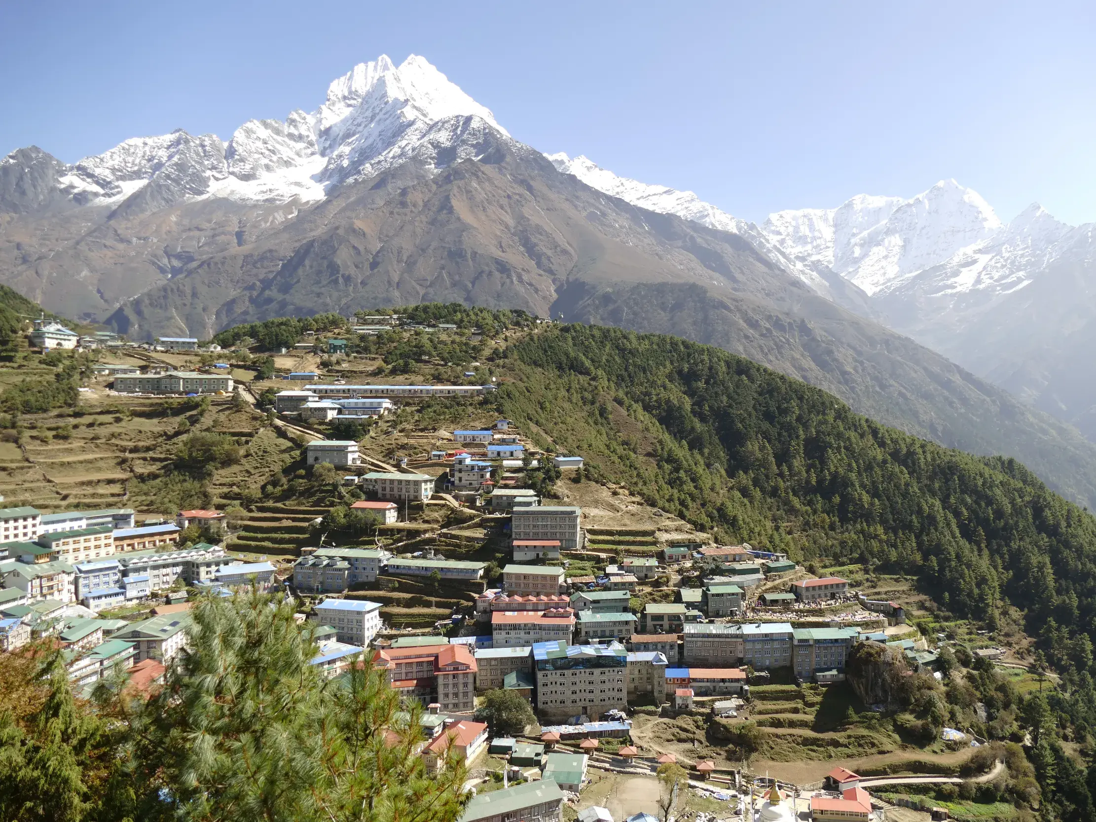 Das Sherpa-Zentrum Namche Bazar an einem terrassierten Berghang im Khumbu-Tal, im Hintergrund schneebedeckte Himalaya-Gipfel.