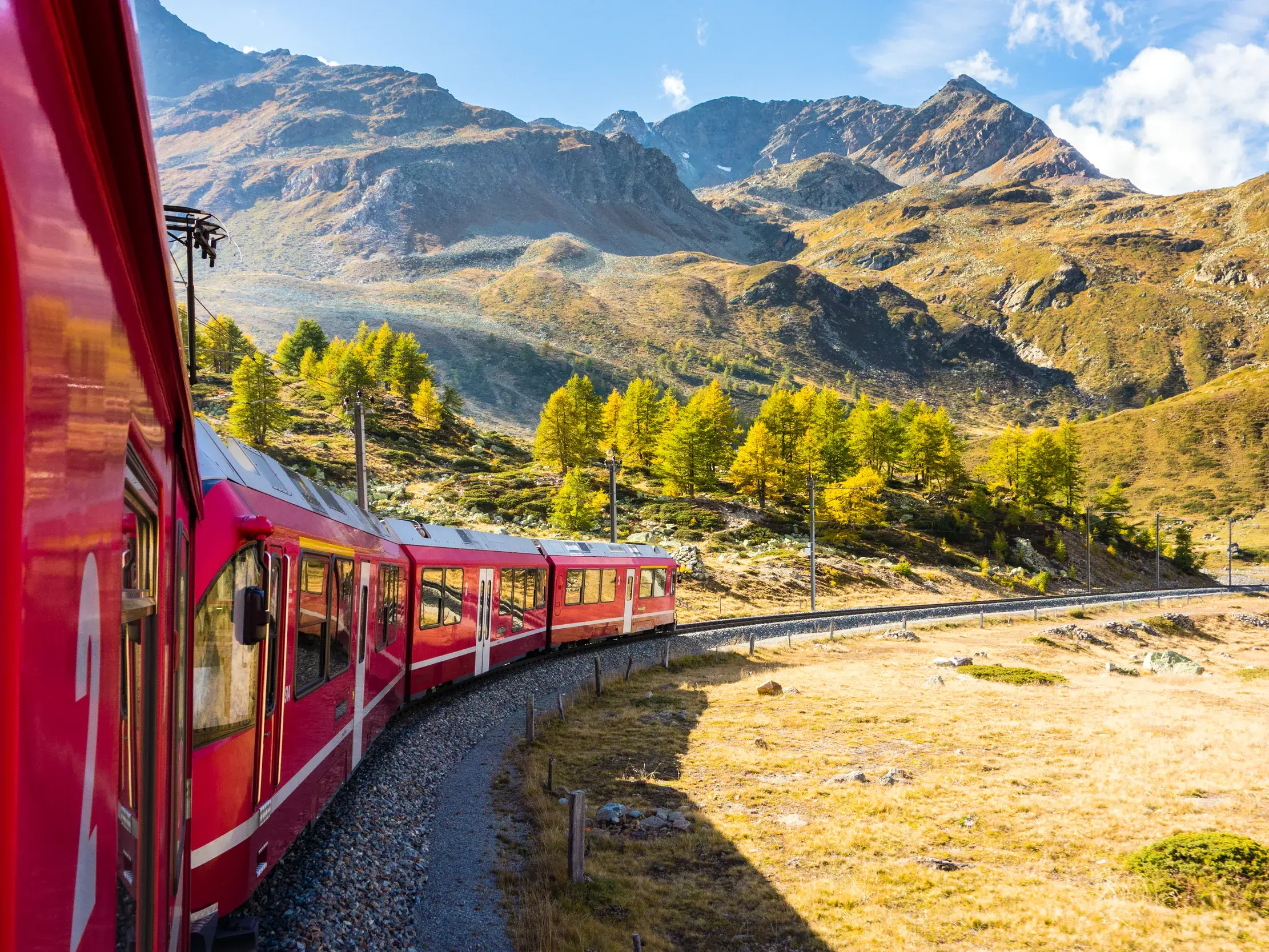 Der rote Bernina Express der Rhätischen Bahn fährt auf einer kurvigen Strecke über den sonnigen Berninapass in Graubünden.