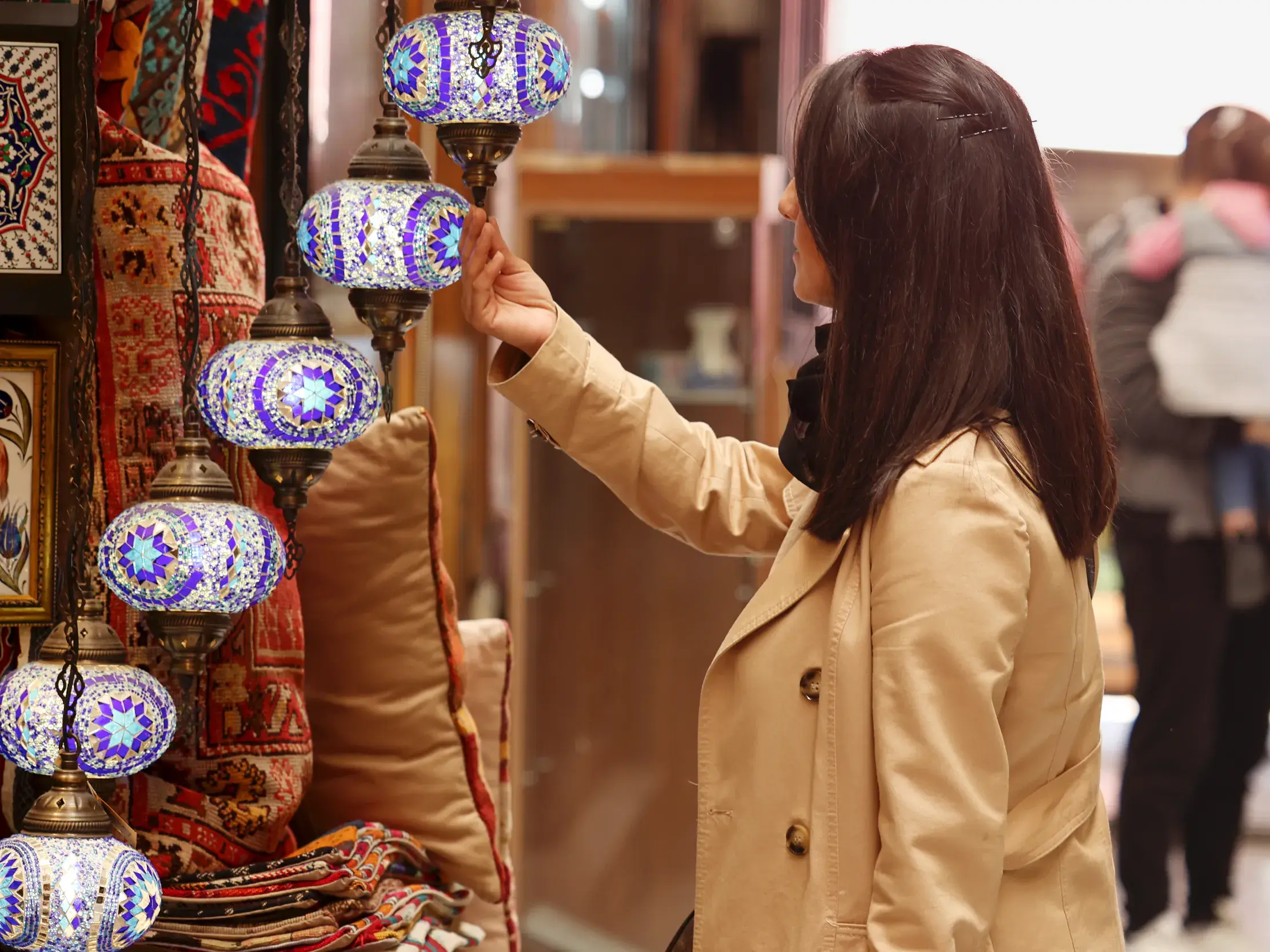 Eine Frau im beigen Mantel betrachtet fasziniert die kunstvollen, blau leuchtenden Mosaiklampen auf einem Basar in Istanbul.