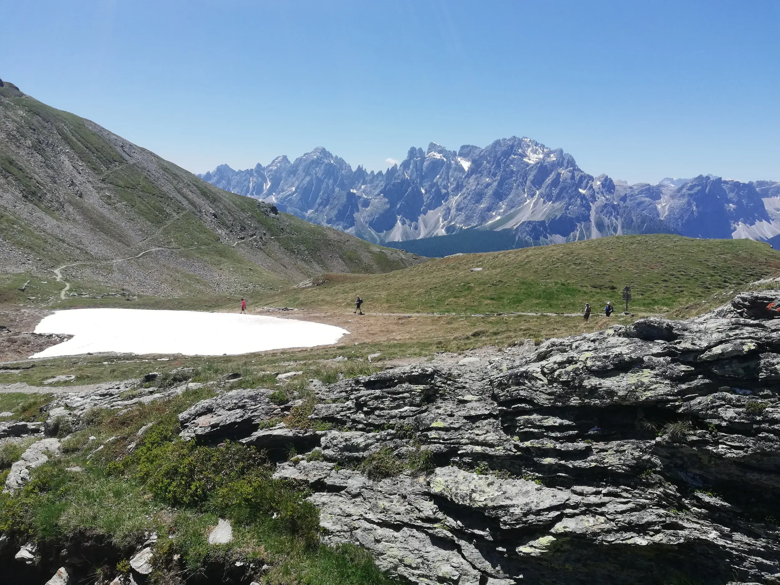 Eine kleine Wandergruppe auf dem Karnischen Höhenweg in den Alpen, vorbei an einem Schneefeld, mit Panoramablick auf die Südtiroler Dolomiten.