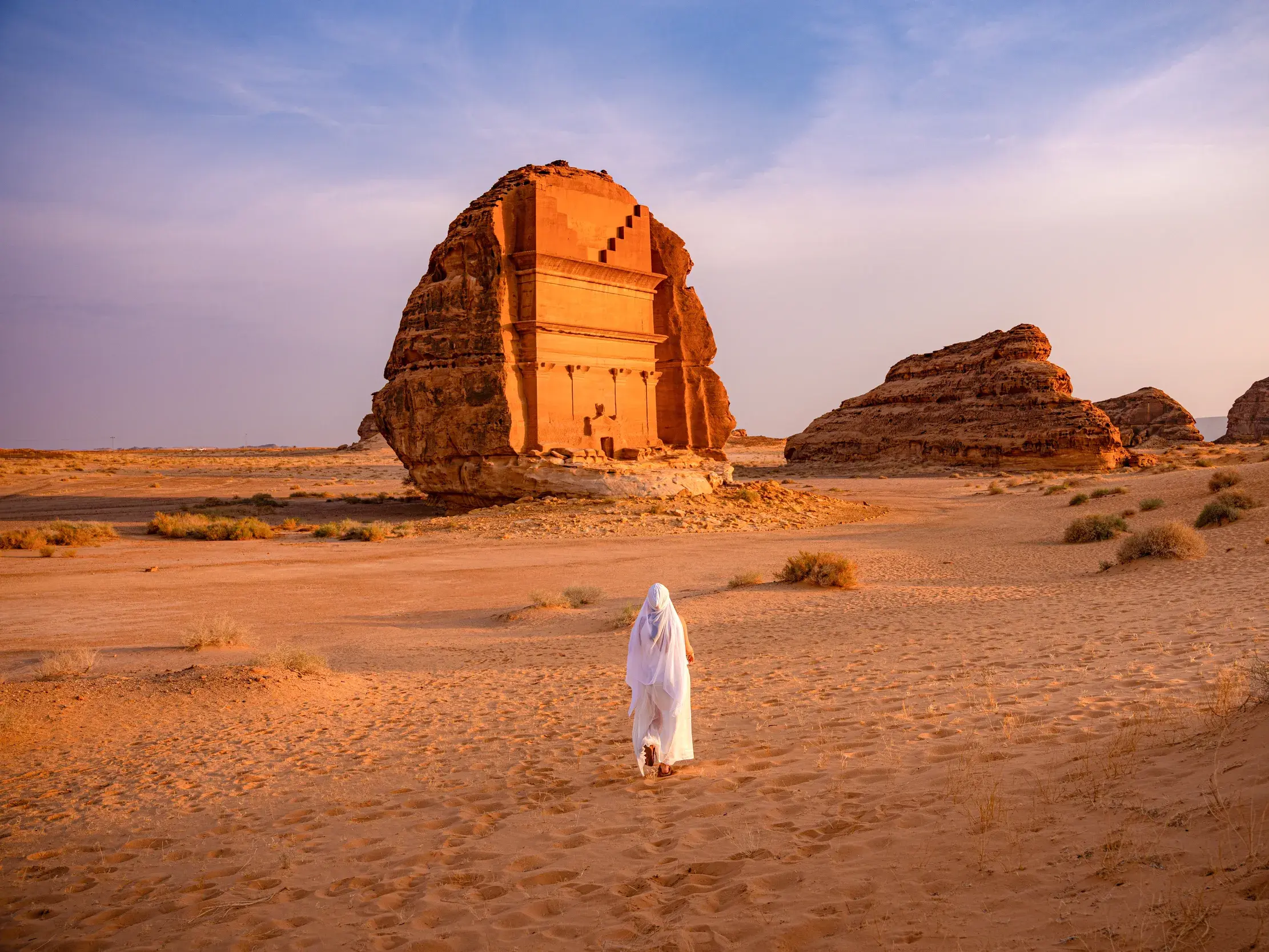 Person in weißem Gewand wandert bei Sonnenuntergang durch die Wüste zum monumentalen Felsengrab Qasr al-Farid in Hegra bei Al-Ula.