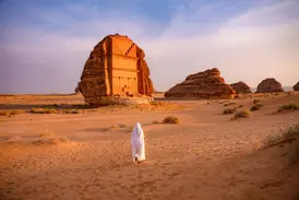 Person in weißem Gewand wandert bei Sonnenuntergang durch die Wüste zum monumentalen Felsengrab Qasr al-Farid in Hegra bei Al-Ula.