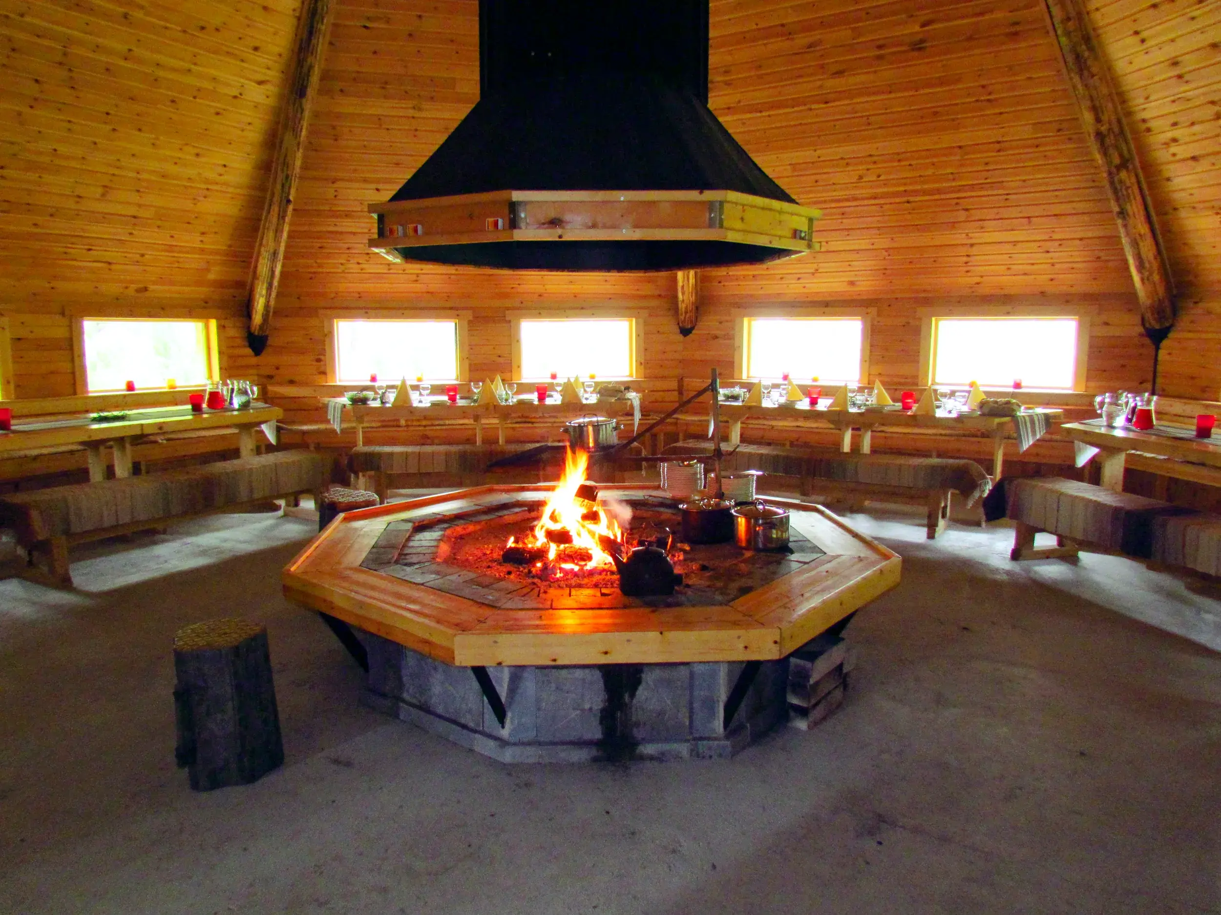 Ein Feuer lodert in der achteckigen Grillhütte der Saija Lodge in Finnland, umgeben von langen, gedeckten Holztischen.