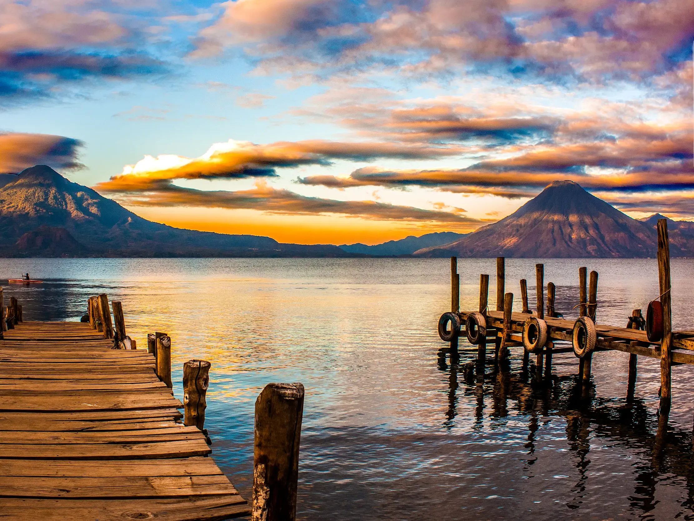 Ein Holzsteg führt am Atitlánsee in Guatemala auf das Wasser, im Hintergrund die Vulkane im Licht des Sonnenuntergangs.