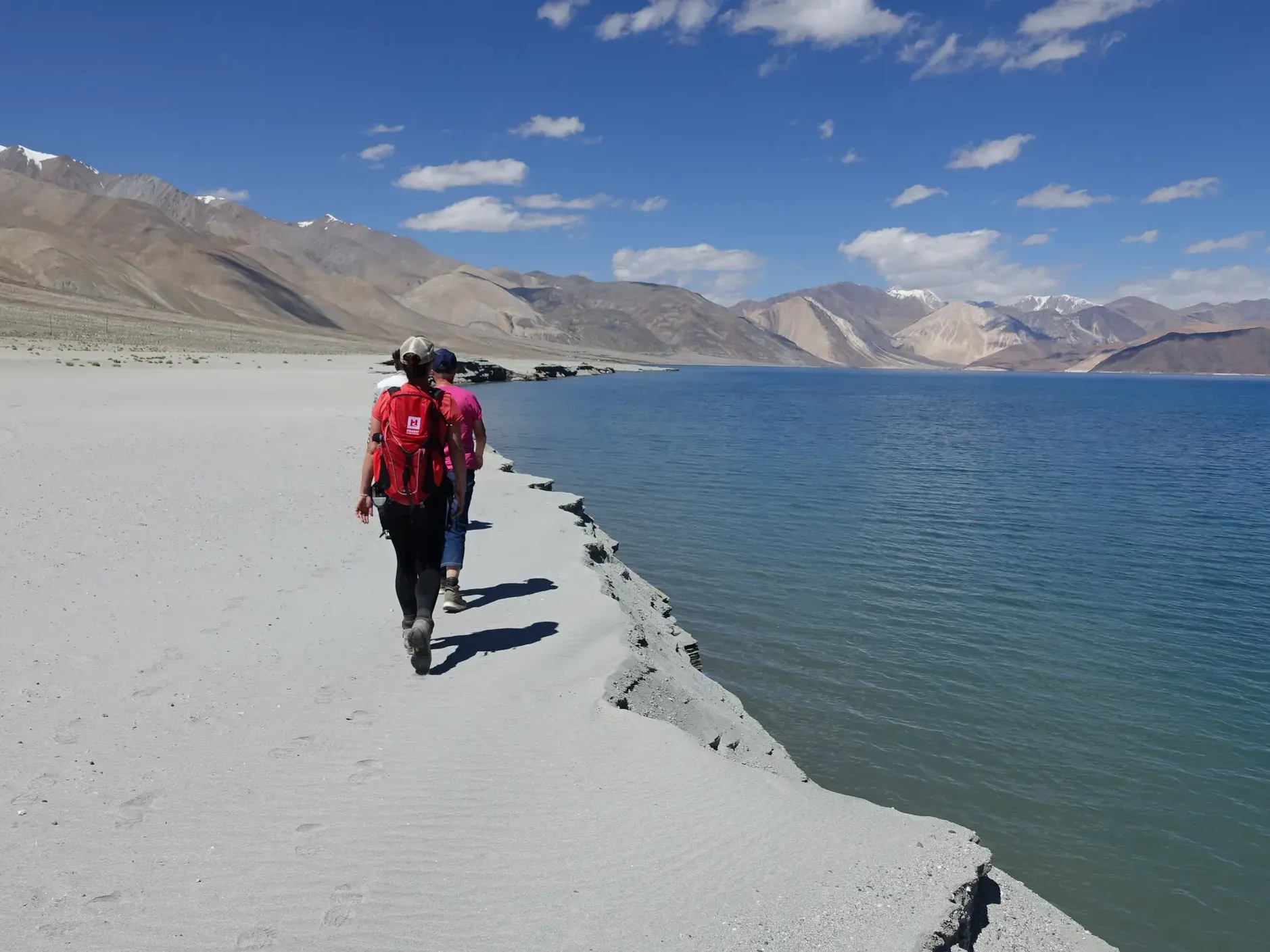 Eine kleine Wandergruppe spaziert am sandigen Ufer des tiefblauen Pangong See in Ladakh vor karger Bergkulisse.