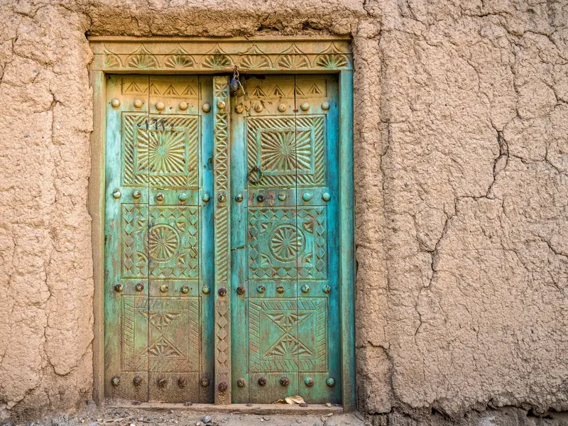 Kunstvoll geschnitzte, türkise Holztür in einer rissigen Lehmfassade eines traditionellen Hauses in Al-Hamra, Oman.