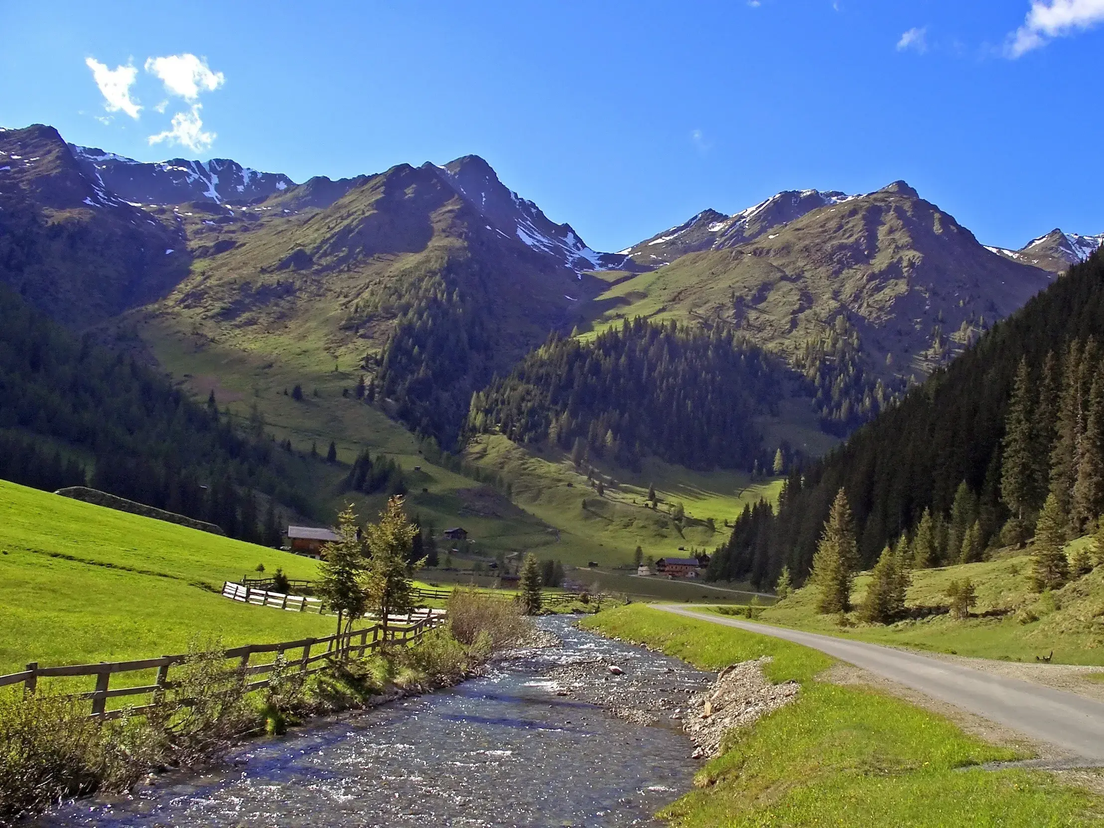 Ein Gebirgsbach fließt durch das sonnige Villgratental in Osttirol, vorbei an grünen Wiesen und schneebedeckten Gipfeln.