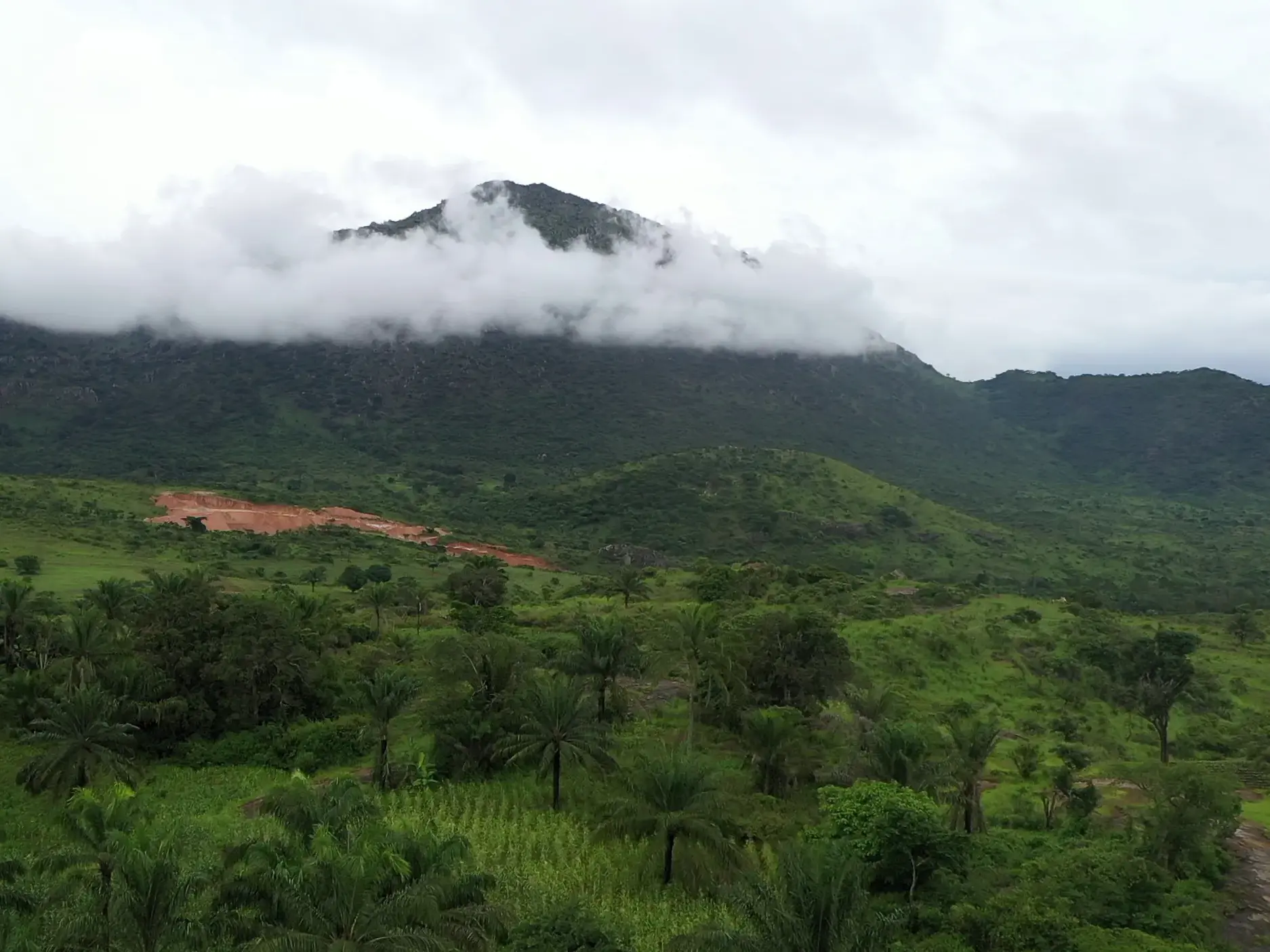Weite, üppig grüne Hügellandschaft des Kumbira Forest Reserve in Angola mit einem wolkenverhangenen Berggipfel im Hintergrund.
