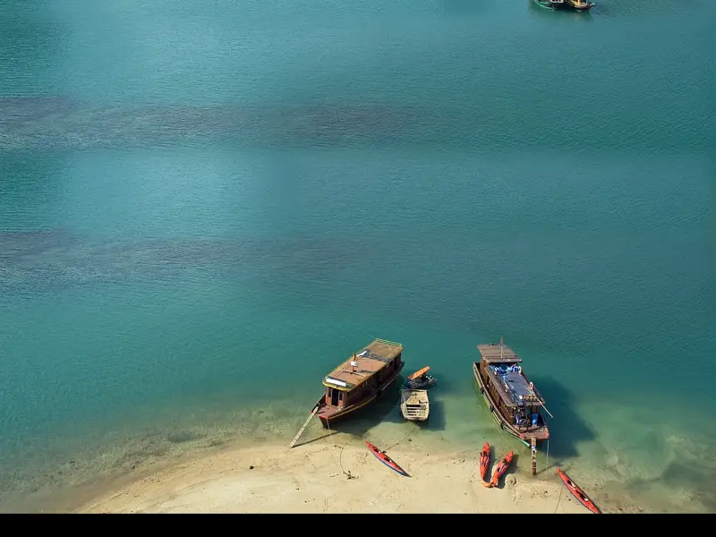 Zwei traditionelle Dschunken und mehrere rote Kajaks liegen an einem Sandstrand im türkisfarbenen Wasser der Halongbucht.