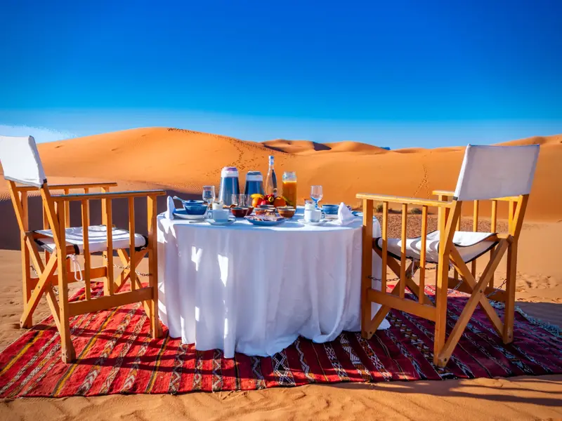 Gedeckter Frühstückstisch für zwei auf einem Teppich inmitten der weiten, orangen Sanddünen von Merzouga in Marokko.