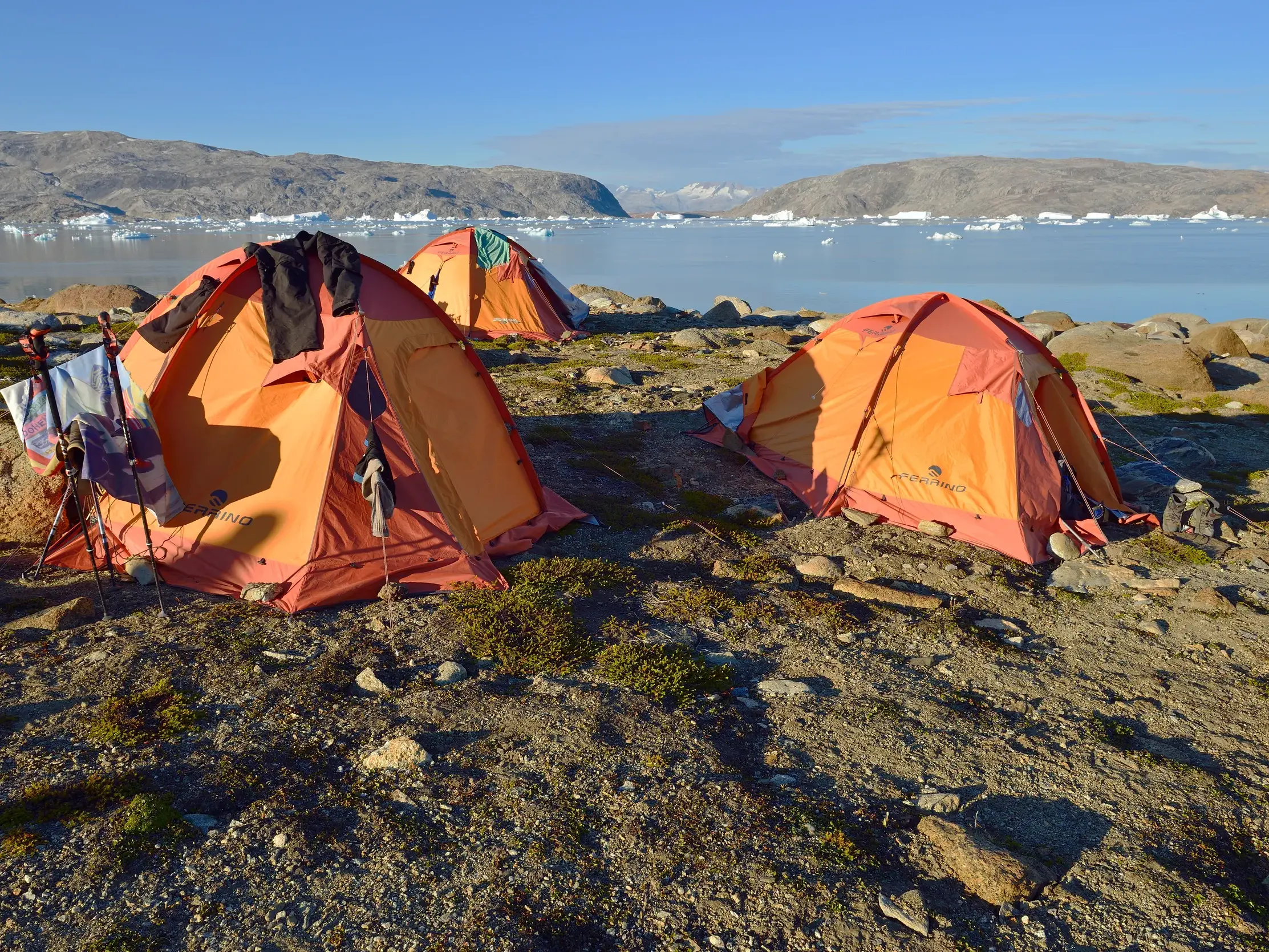 Drei orange Zelte eines Camps am Sermilik-Fjord in Ostgrönland, im Wasser treiben große Eisberge vor Bergkulisse.
