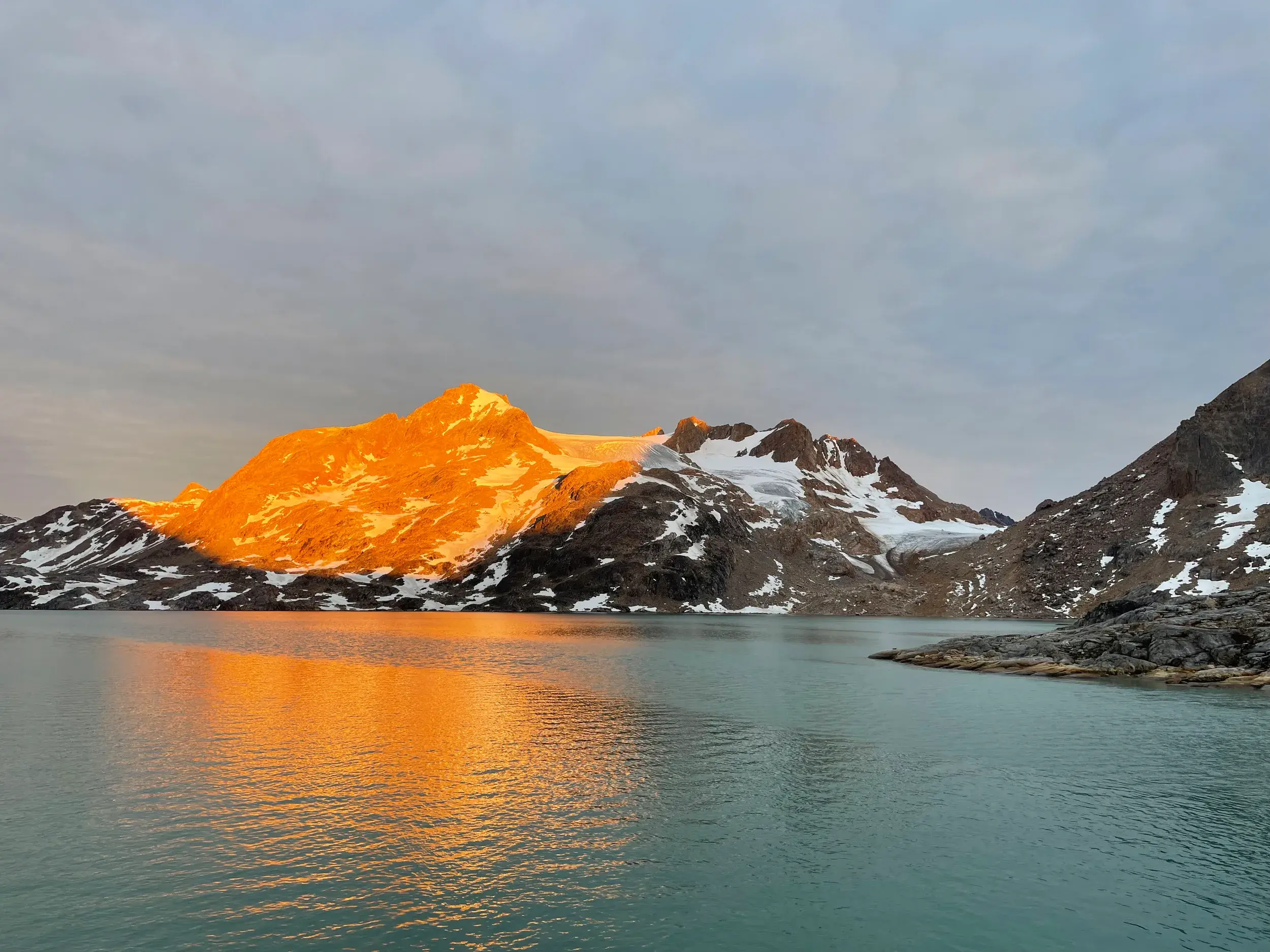 Ein Berggipfel am Sermilik-Fjord in Ostgrönland leuchtet in der goldenen Abendsonne und spiegelt sich im ruhigen Wasser.