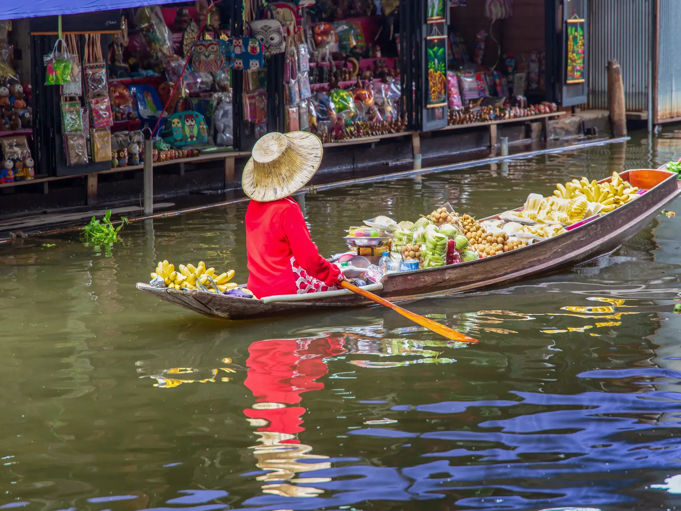 Händlerin mit Strohhut in roter Kleidung paddelt ein mit Bananen und Früchten beladenes Boot auf einem schwimmenden Markt in Thailand.