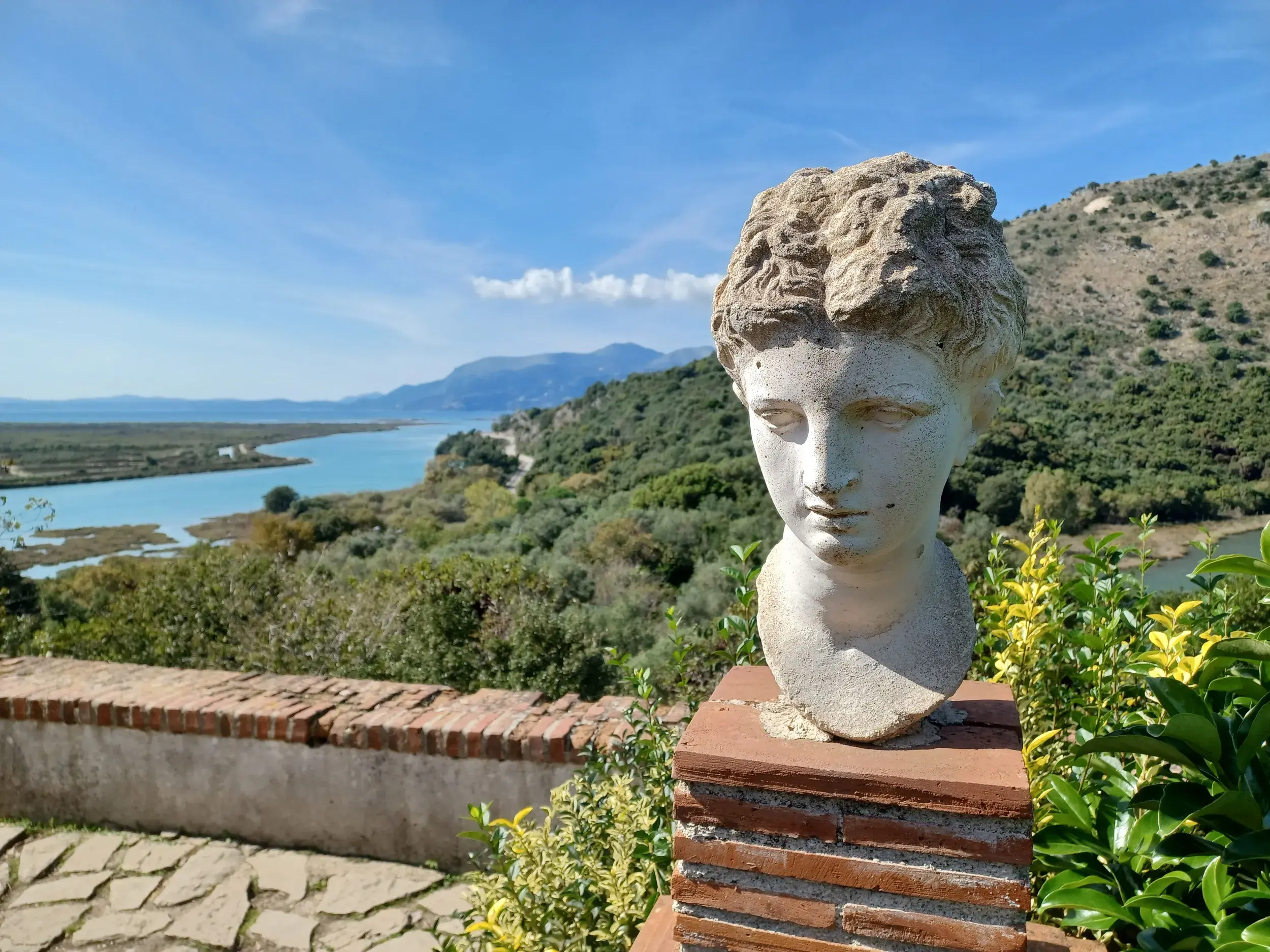 Antike Steinbüste auf einer Mauer im UNESCO-Weltkulturerbe Butrint mit Panoramablick über die Albanische Riviera.