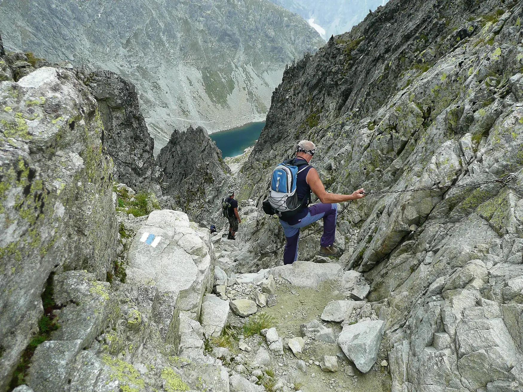 Zwei Wanderer auf einem anspruchsvollen, kettenversicherten Steig in der Hohen Tatra mit weitem Blick auf einen Bergsee.