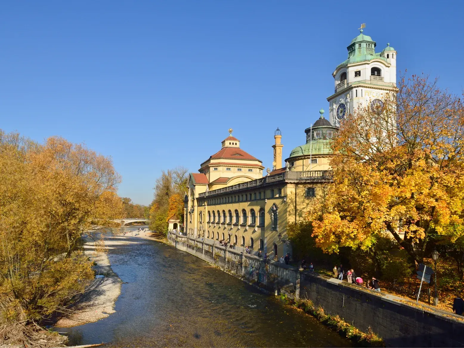Die Isar fließt am Müllerschen Volksbad in München vorbei, umgeben von Bäumen mit goldenem Herbstlaub am blauen Himmel.