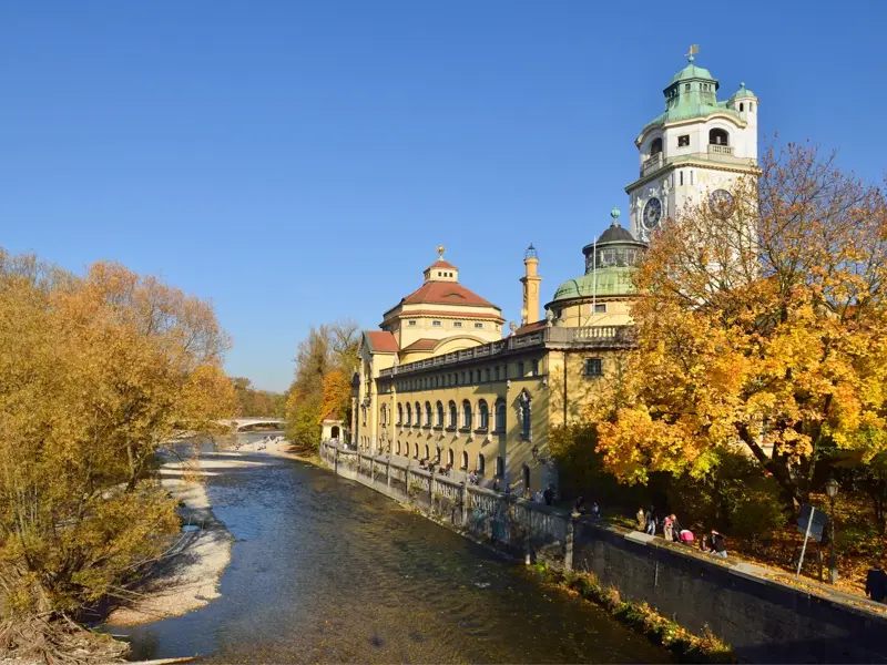 Die Isar fließt am Müllerschen Volksbad in München vorbei, umgeben von Bäumen mit goldenem Herbstlaub am blauen Himmel.