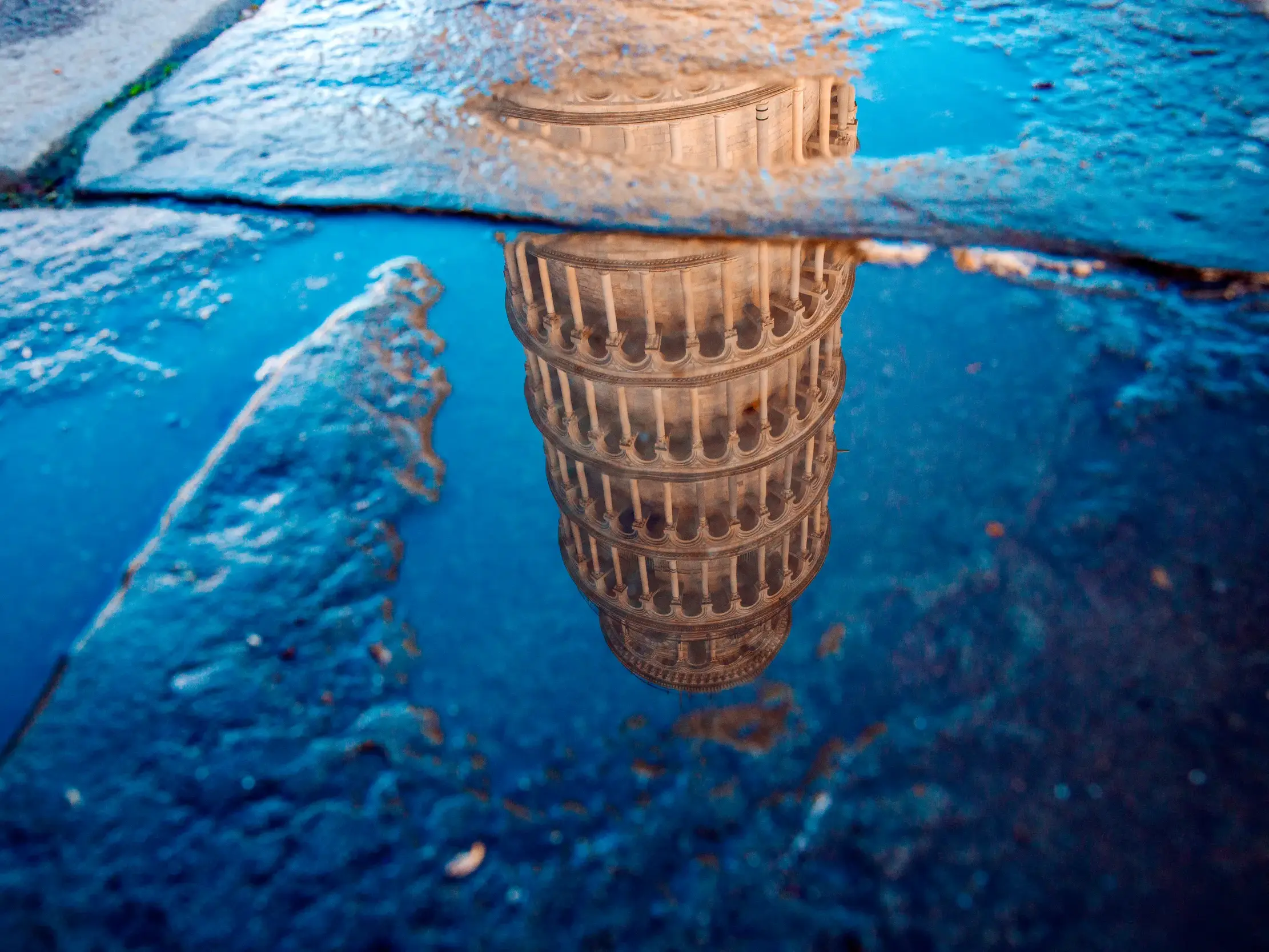 Der Schiefe Turm von Pisa spiegelt sich detailreich in einer blauen Wasserpfütze auf dem Kopfsteinpflaster des Campo dei Miracoli.