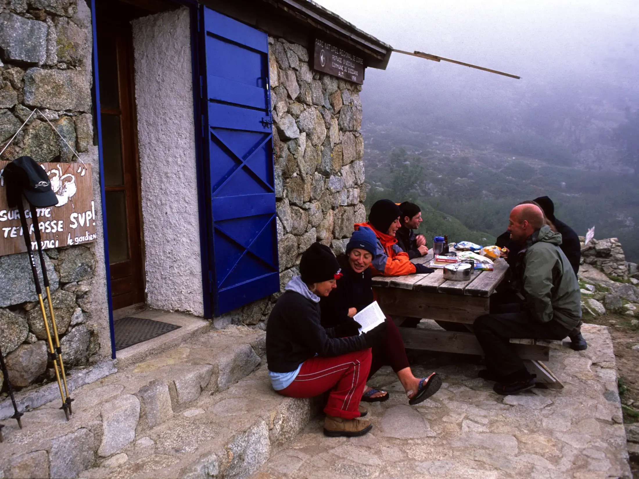 Eine Wandergruppe macht eine gemütliche Pause an einem Holztisch vor einer rustikalen Steinhütte auf dem GR20 in Korsika.