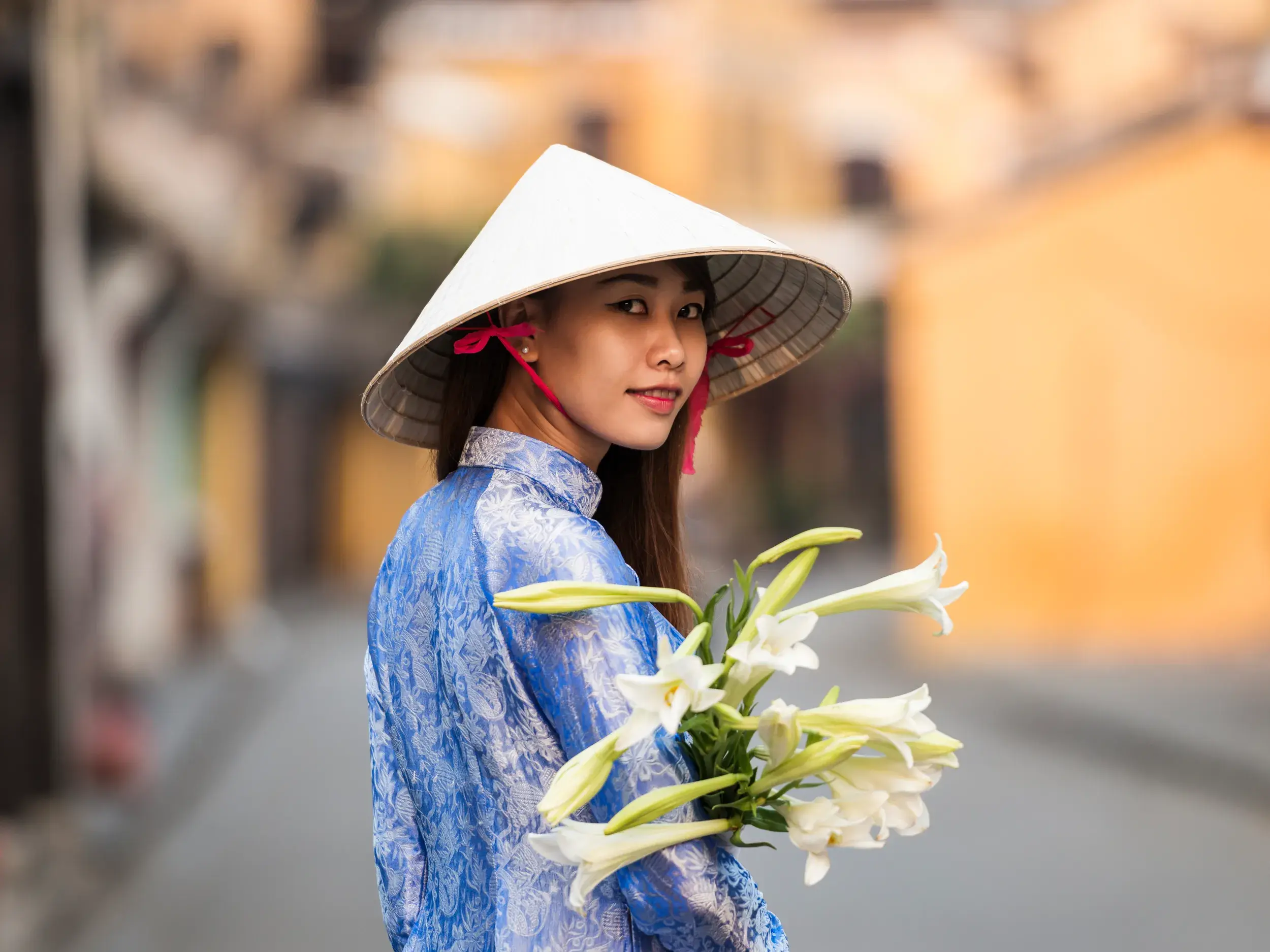 Junge Vietnamesin im traditionellen blauen Ao Dai und Kegelhut mit Lilien in einer Gasse der romantischen Altstadt von Hoi An.