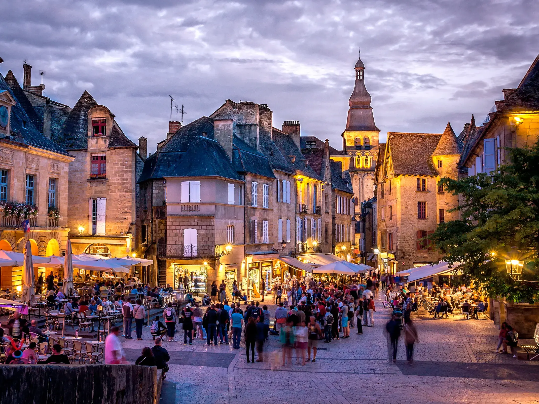 Abendstimmung in Sarlat-la-Canéda: Menschen flanieren über den belebten Platz mit Cafés vor honiggelben Renaissancebauten.