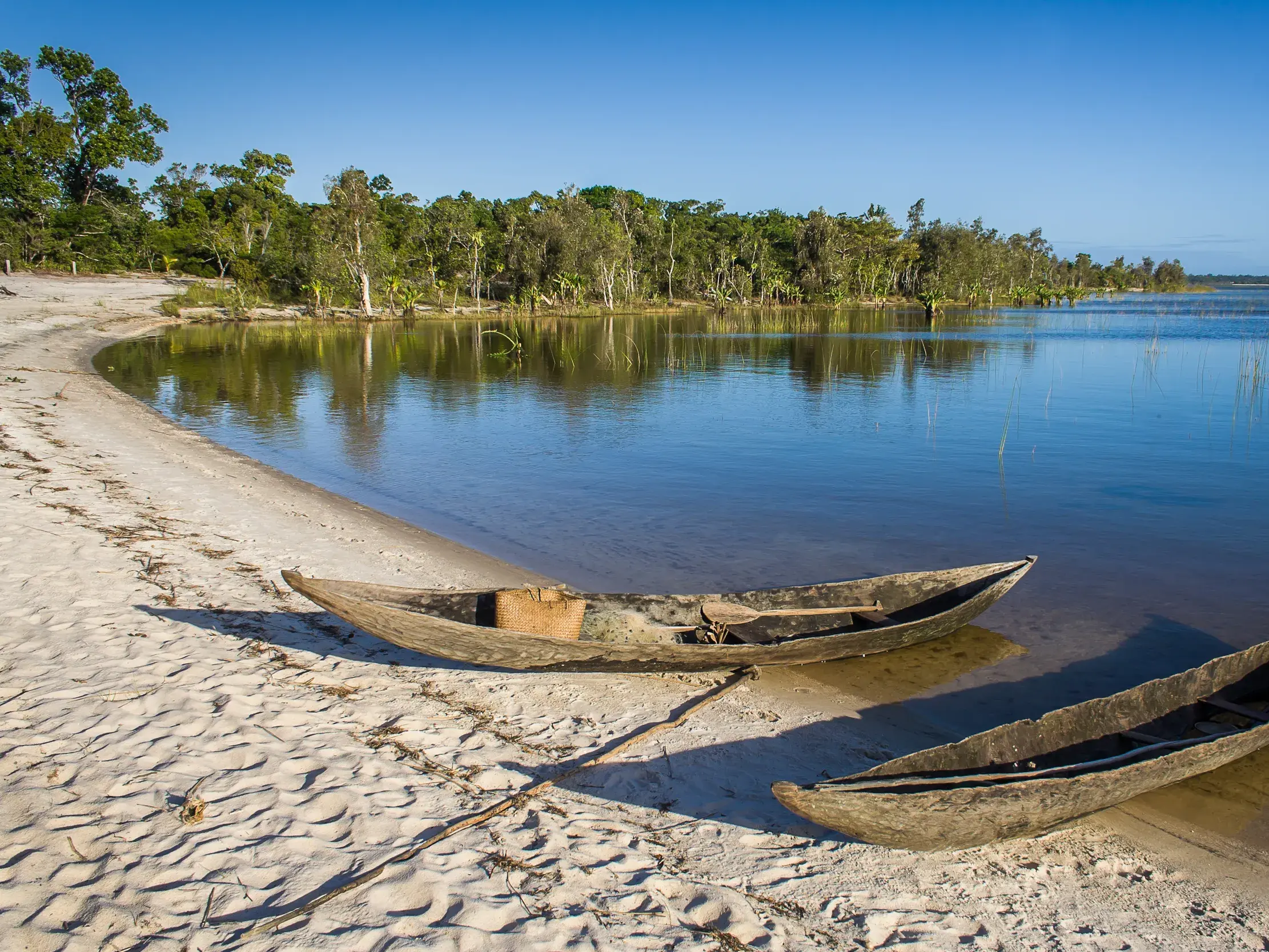 Zwei traditionelle Einbäume liegen am Sandufer des Canal des Pangalanes in Madagaskar, gesäumt von üppigem Regenwald.