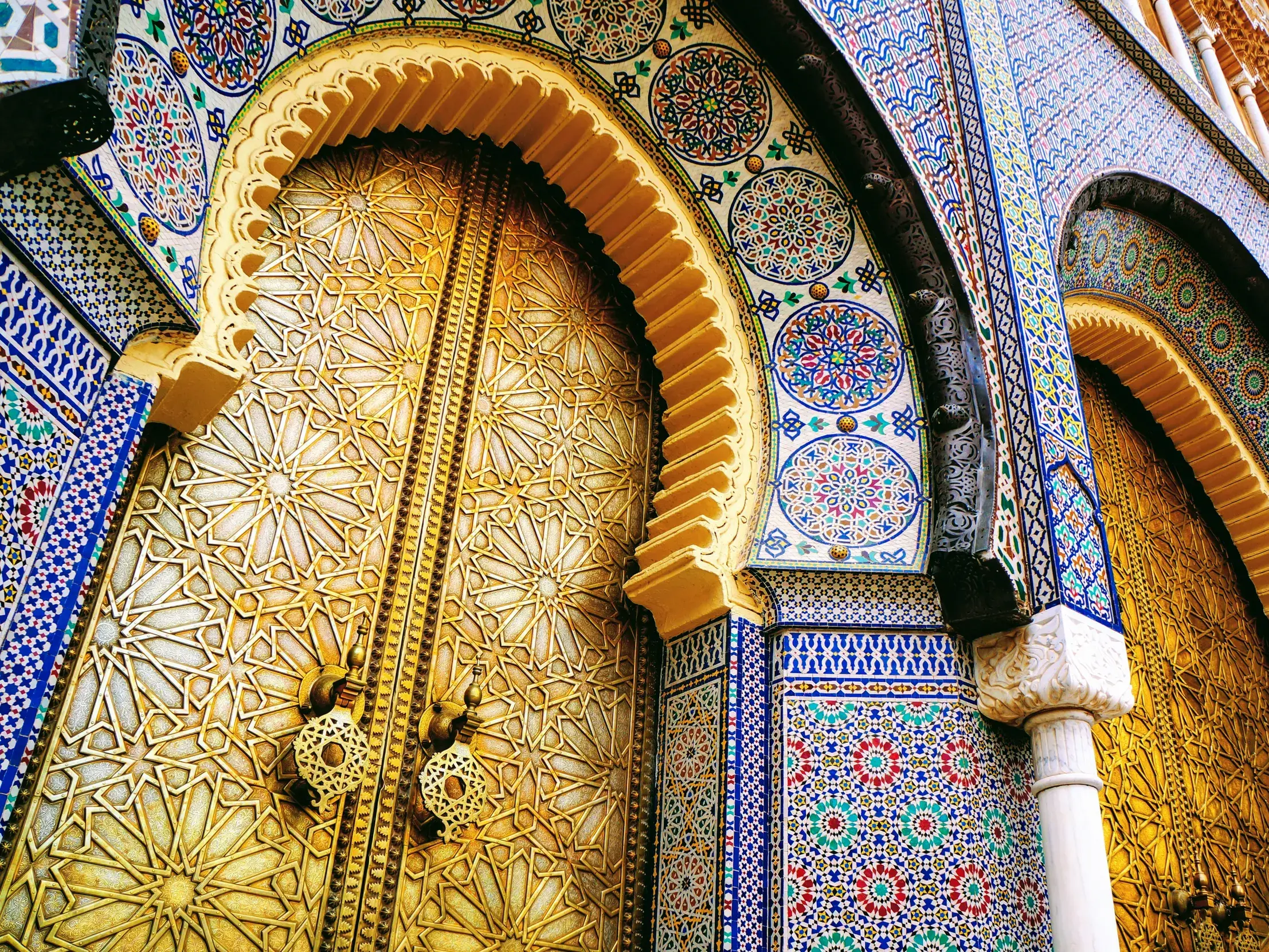 Kunstvoll verzierte, goldene Tore des Königspalasts in Fes, eingerahmt von farbenprächtigen, marokkanischen Mosaikfliesen.