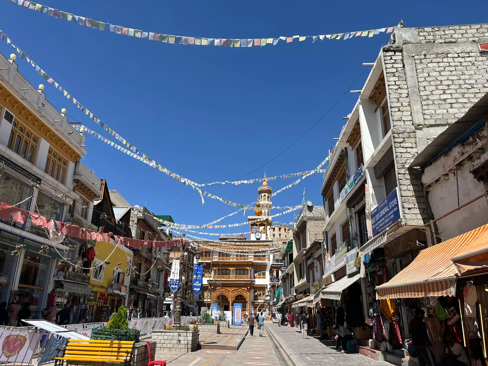 Belebte Gasse am Leh-Markt in Ladakh, gesäumt von Geschäften und überspannt von bunten Gebetsfahnen unter klarem Himmel.
