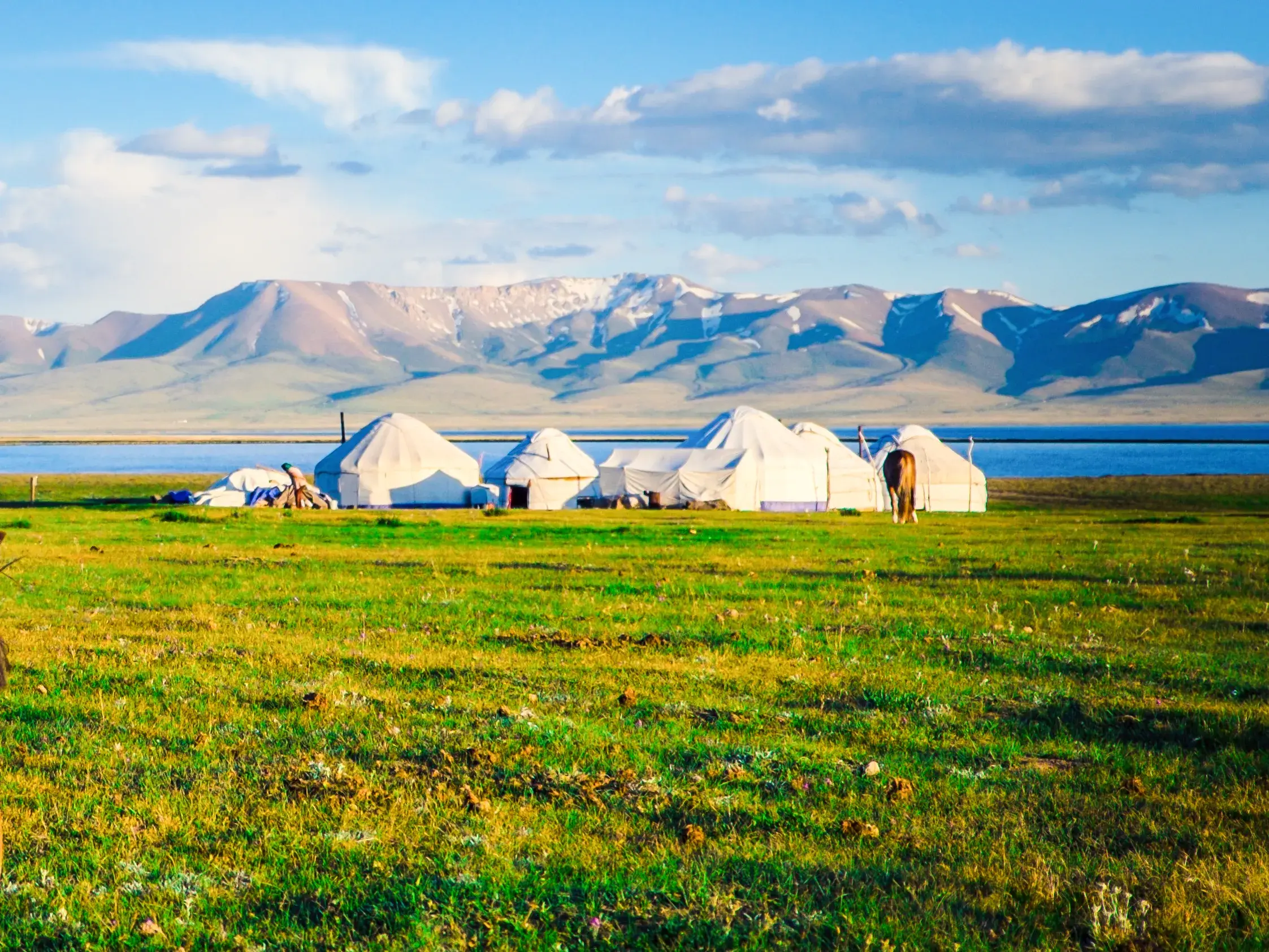 Ein Jurtencamp am Ufer eines Sees vor dem Panorama des schneebedeckten Tianshan-Gebirges in Kirgisistan.